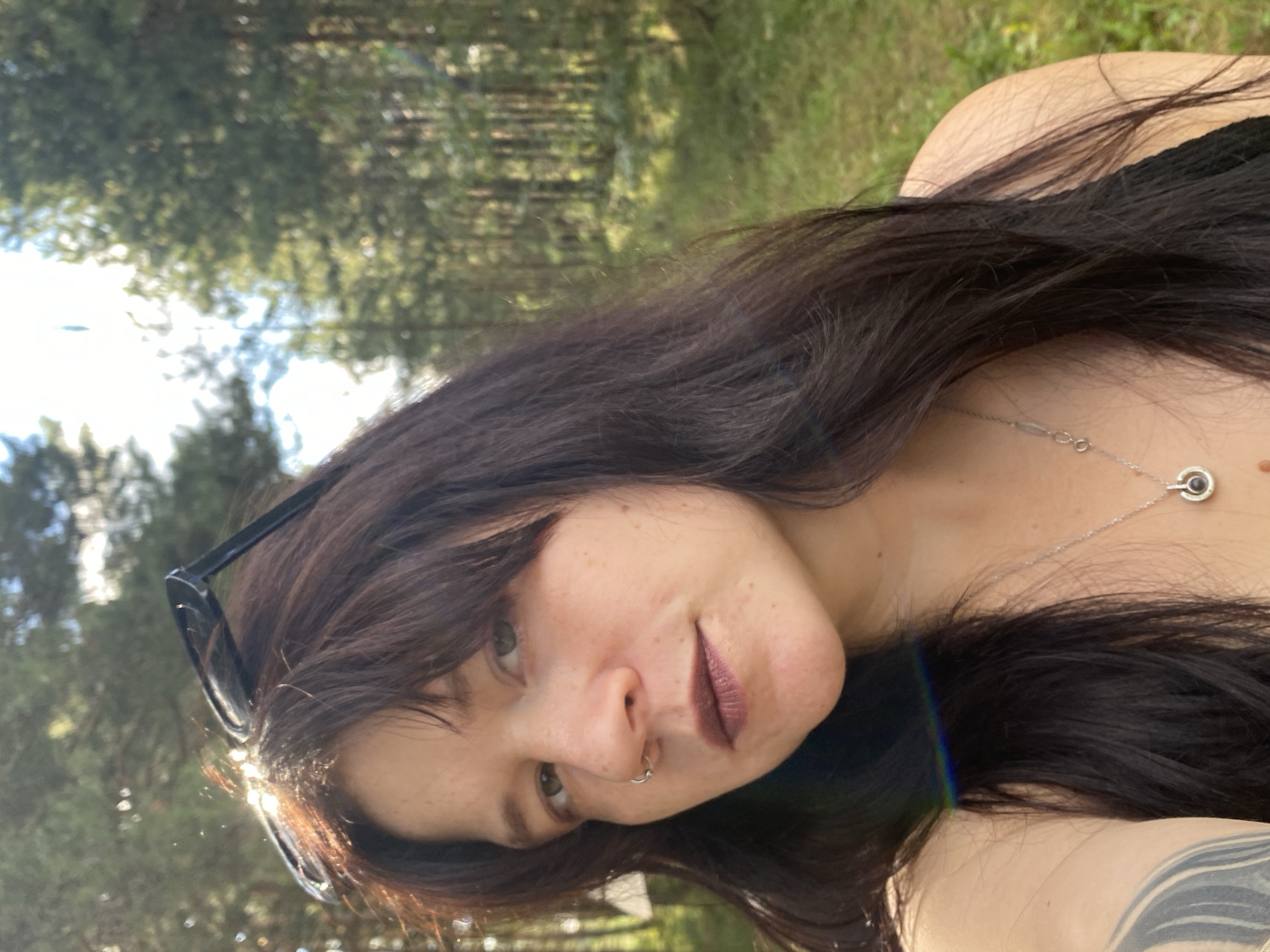 The image is a selfie taken outdoors in a forest setting. A woman with long, dark hair and a nose ring is looking towards the camera. She is wearing a silver necklace with a pendant. Sunglasses are perched on top of her head. The background consists of tall, green pine trees and dappled sunlight, suggesting a natural, possibly rural, environment. There is no indication of the city of Foky, Russia, in the image.