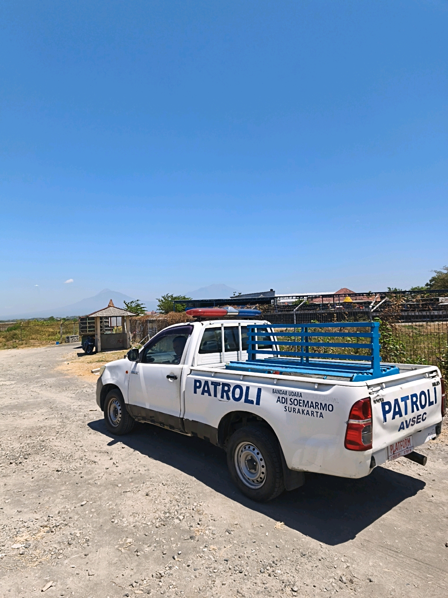 A white patrol truck, marked "PATROLI AVSEC" and "PATROLI" with "BANDAR UDARA ADI SOEMARMO SURAKARTA" on its side, is parked on a dirt road under a clear blue sky. The truck has a blue metal cage in the bed and red and blue flashing lights on its roof. In the background, there is a small open-air structure with a thatched roof, a fence line, distant trees, and mountains. The location is specified as Desa Sindon, Indonesia. The truck is casting a shadow on the ground.