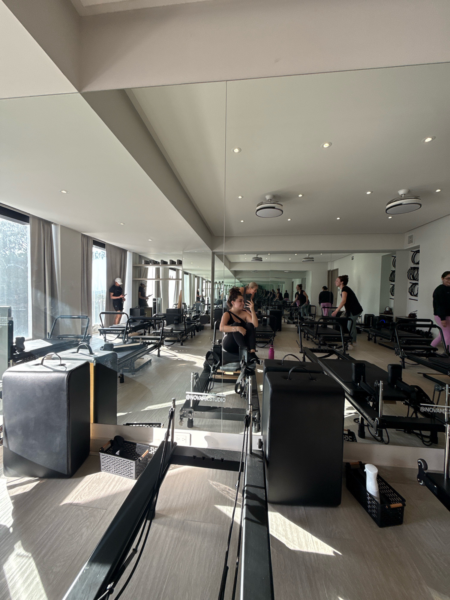 In a well-lit Pilates studio, the scene captures multiple individuals engaged in various activities. The studio features a polished wooden floor and is equipped with numerous Pilates reformers, characterized by their black frames and padded carriages. Large mirrors line one wall, reflecting the studio's layout and activities, creating a sense of spaciousness.
In the foreground, a black Pilates box rests on the floor next to a reformer. A black basket, containing what appears to be workout accessories, is positioned between a reformer and another Pilates box.
In the middle ground, a woman is seated on a reformer, holding her phone and taking a selfie, wearing a black tank top and leggings. Nearby, another woman is standing, appearing to give instructions or assistance. Further back, several other individuals are visible, some standing and others on reformers, also participating in exercises. A man in a black t-shirt and white cap stands near a window, looking at his phone.
The background reveals more Pilates reformers, shelves with equipment, and the continuation of the mirrored wall. Natural light streams in through large windows covered by light grey curtains, casting distinct shadows across the floor, indicating it is daytime. The ceiling is white and features recessed lighting and modern circular ceiling fans. The overall environment is clean, organized, and suggests an active fitness session. The specific location of San Bernardino Tlaxcalancingo, Mexico, suggests a modern, potentially upscale fitness facility within that community.