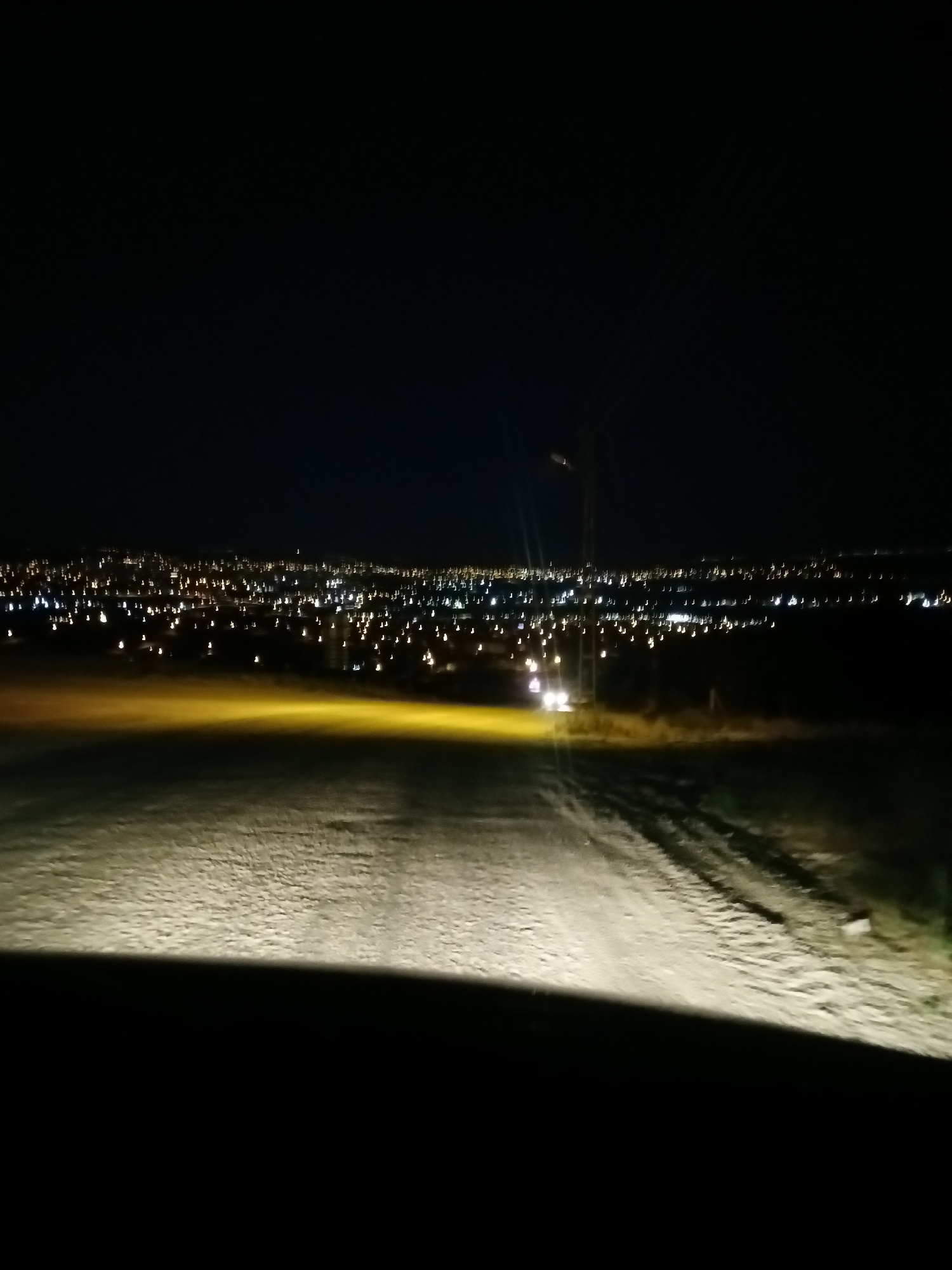 The image shows a dark landscape with a distant city illuminated by numerous lights. The foreground is a dirt road, likely illuminated by the headlights of a vehicle not fully visible, casting a bright, yellowish light. The road has visible tire tracks and a textured surface. Beyond the road, there is a field or undeveloped land. The middle ground features a prominent, thin vertical pole with a light fixture, a common sight in urban or suburban areas, and two bright white lights that appear to be from a vehicle driving on the road. The background is dominated by the sprawling city lights, which appear somewhat blurred, suggesting either distance, motion, or a shallow depth of field. The city lights stretch across the horizon, indicating a significant urban area. The sky above is completely black, devoid of stars or moon, indicating nighttime. The overall impression is that of a view from a vehicle approaching or overlooking a city at night from a slightly elevated position. The city is likely Bahşılı Belediyesi, Turkey, given the context.