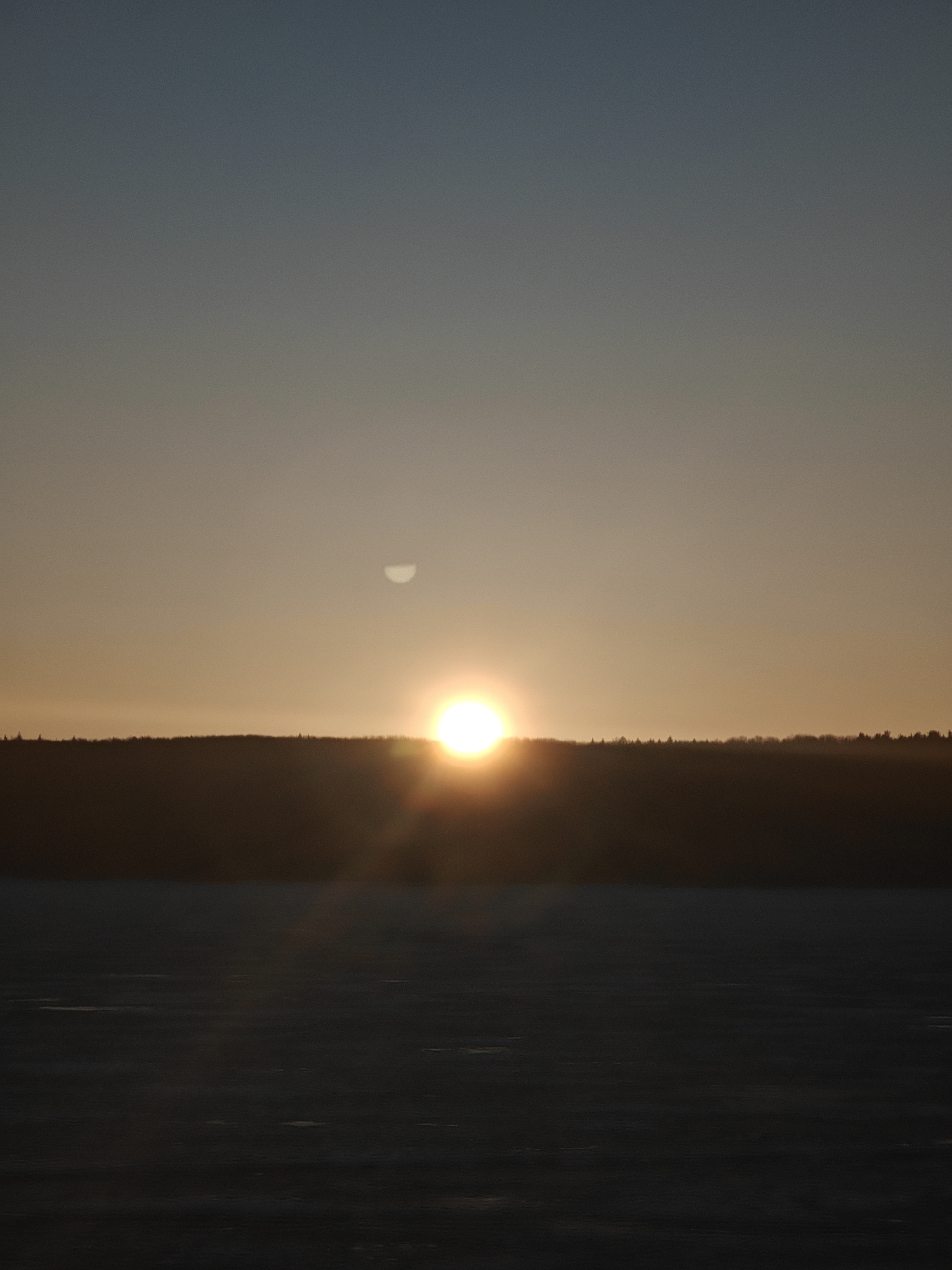 The image captures a low sun, partly obscured by the horizon. A distinct, semi-circular shape, possibly the moon, is visible above the horizon line. The foreground shows a dark, possibly frozen, body of water. In the middle ground, a dark, horizontal band likely represents a forest or tree line. The sky transitions from a dark blue/gray at the top to a yellow-orange near the horizon. The location is identified as Chaikovsky, Russia.
