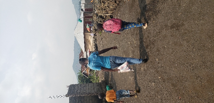 In Buzove, Democratic Republic of the Congo, three individuals walk away from the viewer on an unpaved, gravel-strewn dirt path. The central figure is an adult male wearing a light blue polo shirt and blue jeans, carrying a white plastic bag in his left hand. To his left, a child in an orange t-shirt and blue jeans walks ahead. To his right, a child wearing a pink backpack and patterned blue pants walks alongside. The path is bordered by a partially constructed stone block wall on the left and a low stone wall on the right. In the mid-ground, several single-story structures with various roof types are visible, including one with a light-colored corrugated metal roof and wooden walls. Distant mountains are visible under a light grey, overcast sky.