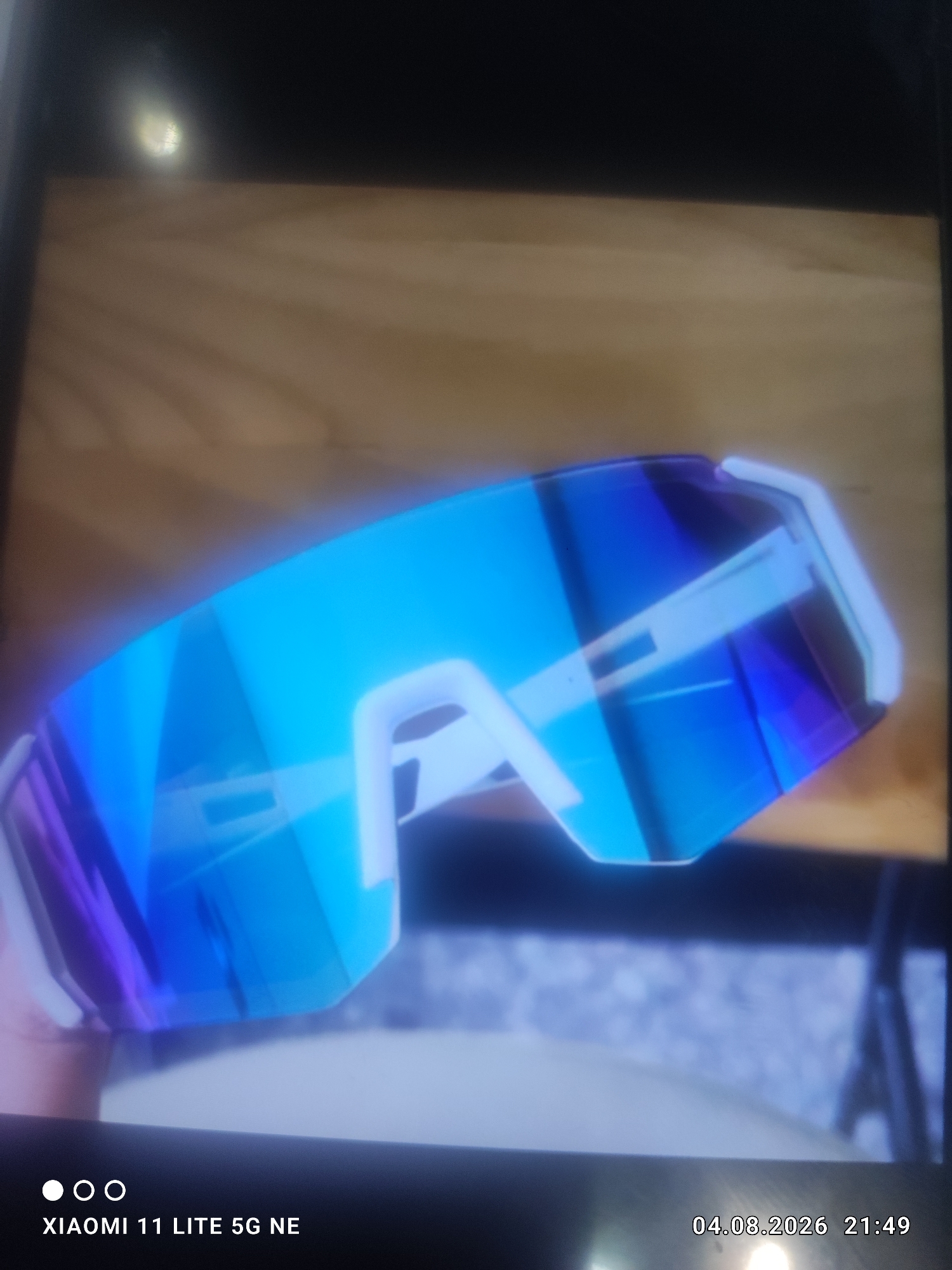 A pair of futuristic, blue and purple mirrored wrap-around sunglasses is displayed on a light brown wooden surface. The sunglasses have a white frame with a thick bridge and white accents on the arms. The lenses are highly reflective, showing a blend of bright blue and deep purple hues. The background is blurred, with hints of dark and light shades, suggesting an indoor or shaded outdoor environment. The timestamp "04.08.2026 21:49" and "XIAOMI 11 LITE 5G NE" are visible in the lower right and left corners respectively. There is no information about the location in Kara-Chungur, Kyrgyzstan within the image.