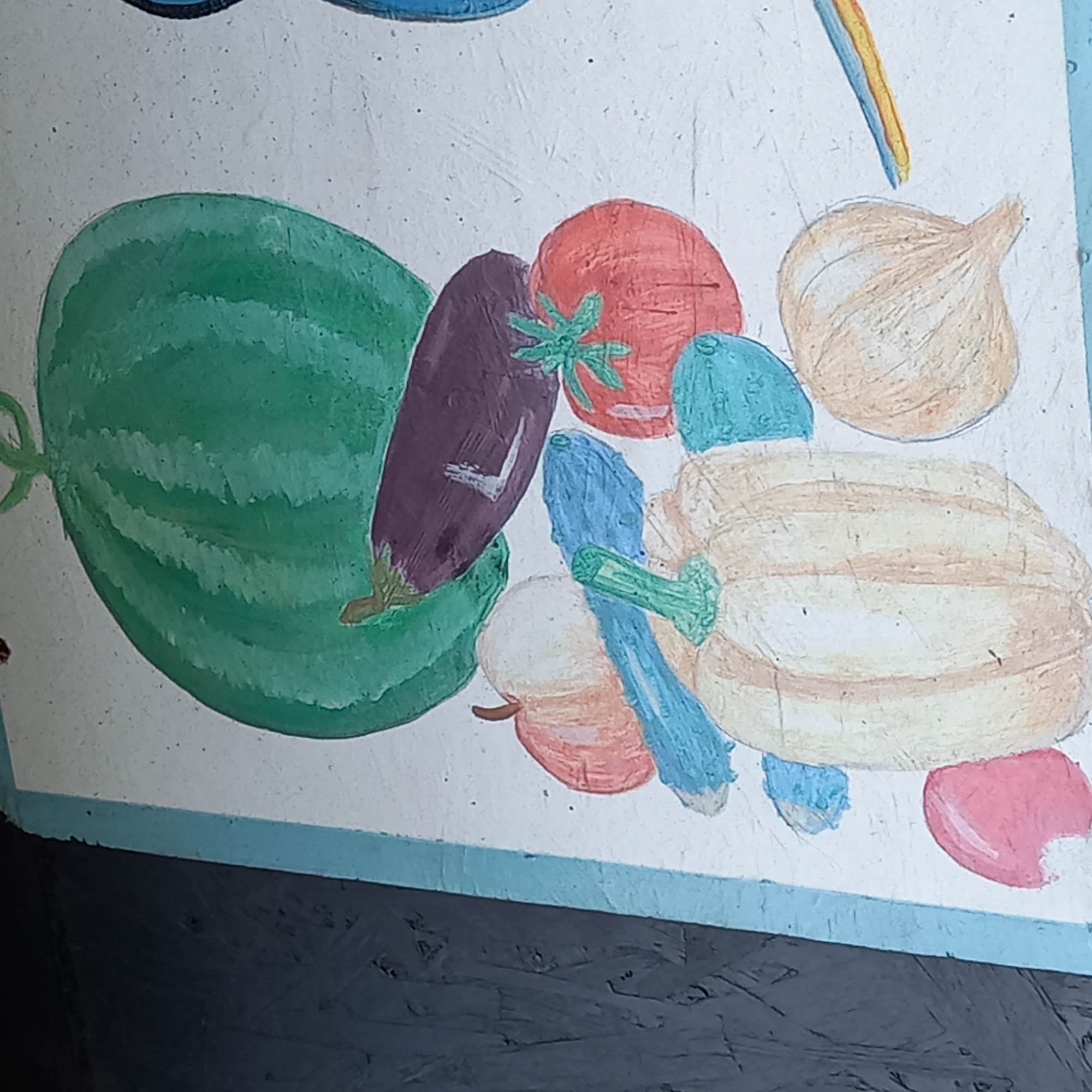 This image displays a section of a painted mural or sign, likely located in Velykodolynske, Ukraine. The artwork depicts a collection of fruits and vegetables in a folk-art style against a white background.
Prominently featured are:
* A large, green watermelon with darker stripes, positioned at the top.
* A dark purple eggplant situated diagonally below the watermelon.
* A red tomato with green foliage on its stem, located to the right.
* A light tan, vertically striped pumpkin or squash with a green stem, positioned centrally at the bottom.
* An orange-red apple with a brown stem, to the left of the pumpkin.
* An elongated blue vegetable, possibly a cucumber or zucchini, placed diagonally behind the apple and pumpkin.
* A light tan, bulbous onion or garlic clove with a pointed end, on the bottom right.
* Several smaller, irregular blue shapes are scattered around the main produce items.
* A fragmented pink object is visible on the bottom left.
* A thin, multi-colored strip (blue, yellow, orange) extends from the right edge.
The painting is executed on a textured white surface, possibly wood or plaster. A light blue painted border separates the artwork from a dark, textured, possibly wooden or concrete surface on the far left of the frame. The overall impression is a simple, vibrant representation of agricultural produce. There are no people present in the scene.