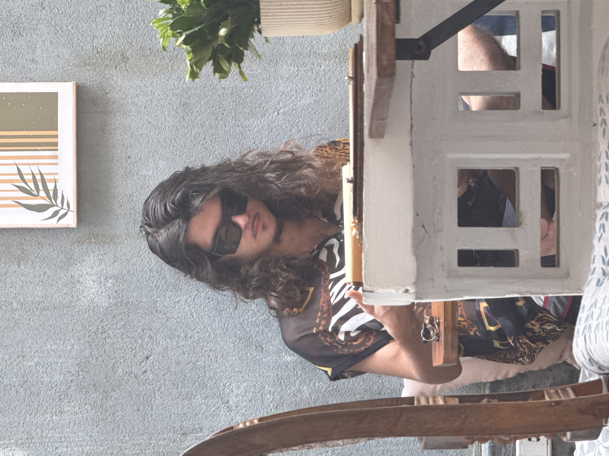 A medium shot depicts a man in sunglasses, with long, wavy dark hair, wearing a black and brown patterned shirt. He is looking to the left, obscured in part by a white, multi-paned, window-like object. To the left of the man, there is a wall with a minimalist painting. Above the man, in the upper portion of the frame, is a potted plant. In the bottom foreground is a wooden object, possibly a chair. The setting appears to be Đà Nẵng, Vietnam. The scene suggests an indoor or semi-outdoor space, possibly a café or home interior.