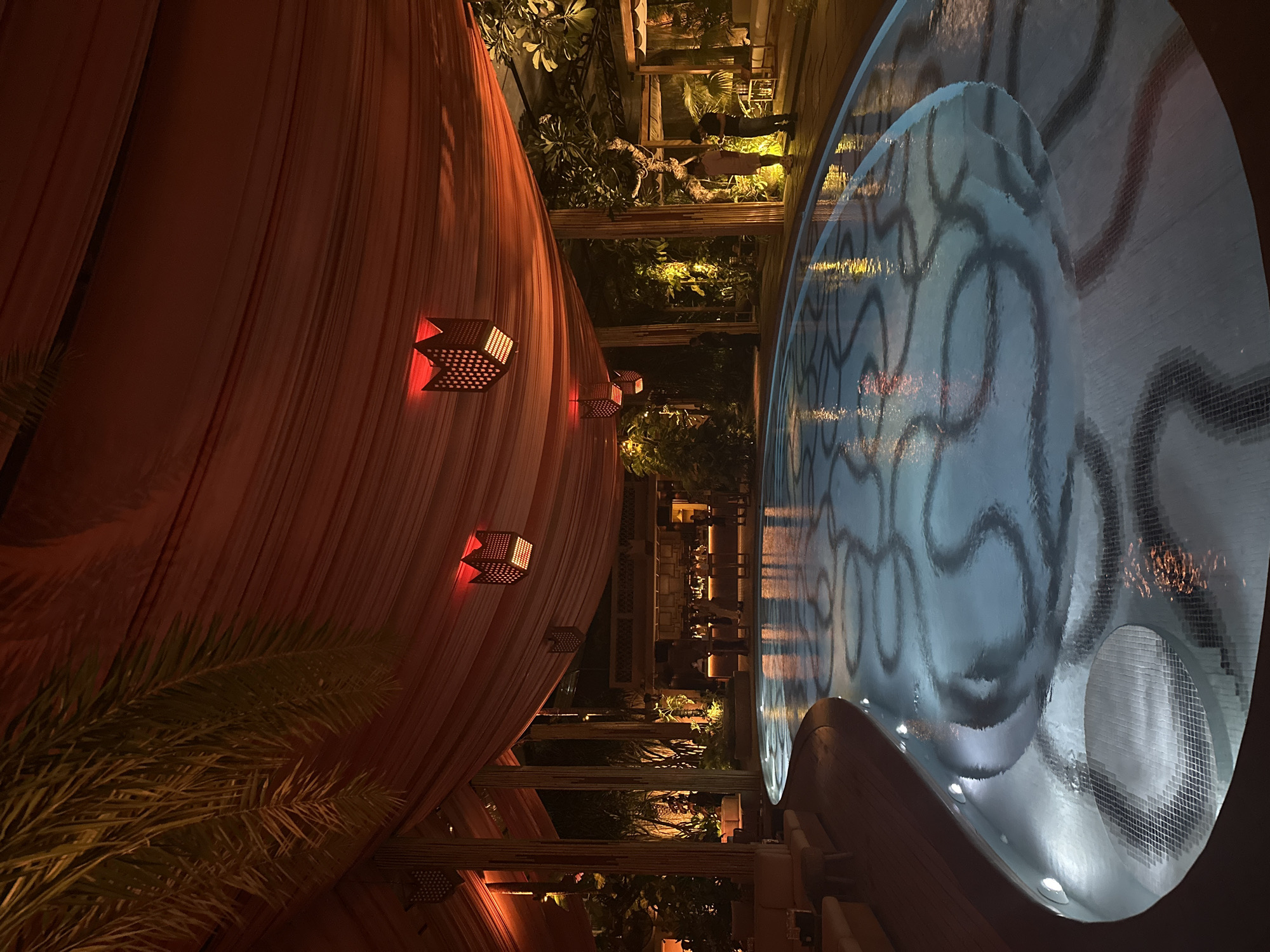 The image shows an outdoor event space at night. A long, draping orange fabric hangs from the left side, adorned with small, cube-shaped lanterns emitting a warm red light. To the right, a curved water feature with a patterned surface reflects the surrounding lights. In the background, a bar area with stools and people can be seen, as well as garden foliage. The location is North Kuta, Indonesia.