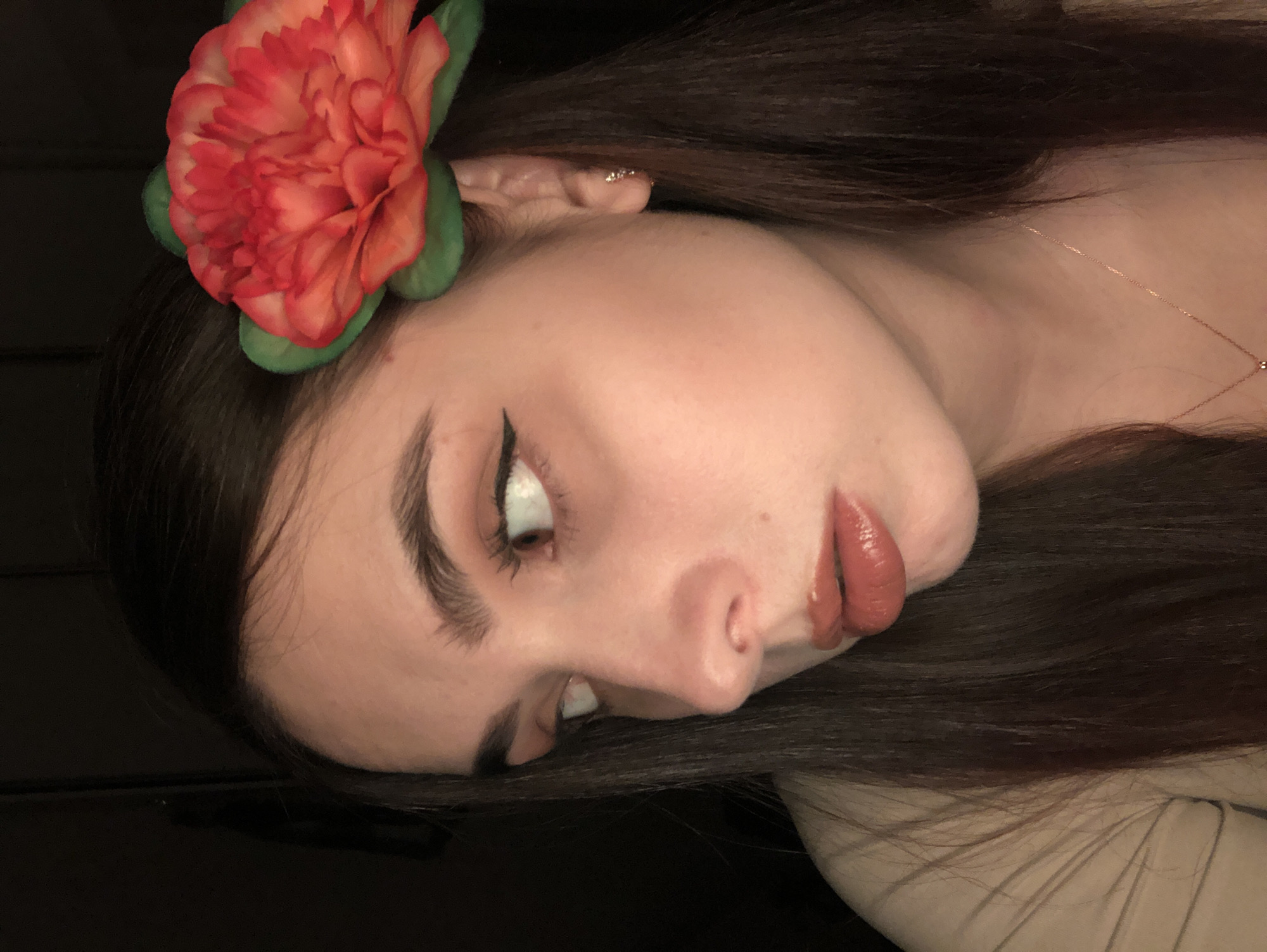 A close-up image depicts a woman's face in profile, tilted to the right. She has dark hair and fair skin. A large, artificial red flower is positioned near her ear. She is wearing eyeliner and lipstick. A delicate necklace is visible on her neck. The background is dark and indistinct. The image does not provide any information to indicate the location is Noviy, Russia.