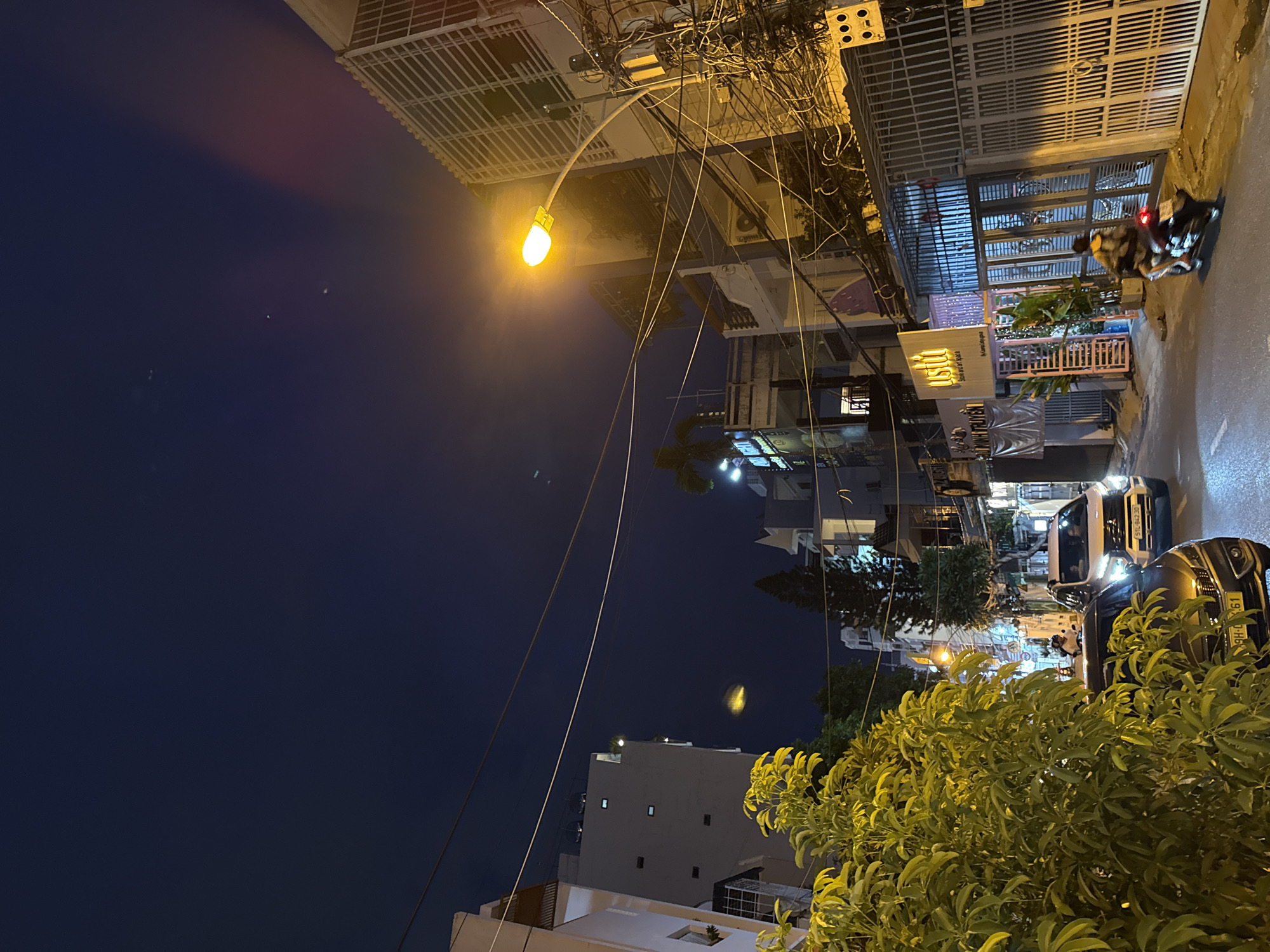 This is a nighttime street scene in Nha Trang, Vietnam. The image captures a narrow street lined with buildings, many of which appear to be residential or commercial establishments. A prominent street lamp illuminates a portion of the road, casting a warm glow on the surroundings.
Several vehicles are visible, including a white SUV and a dark-colored car parked on the street. A person is riding a motorcycle, traveling down the street. The buildings display various architectural styles, with some featuring balconies and intricate metalwork. Overhead, a complex network of electrical wires is strung across the street, typical of urban environments in Vietnam.
The sky is dark blue, indicating it is either late evening or early morning, with a few faint stars or distant lights visible. The presence of lush green foliage from a large tree in the foreground suggests a tropical climate. Signage in Vietnamese can be seen on some of the buildings, indicating businesses or residences. The overall impression is of a bustling urban area at night, with the characteristic sights and sounds of street life.