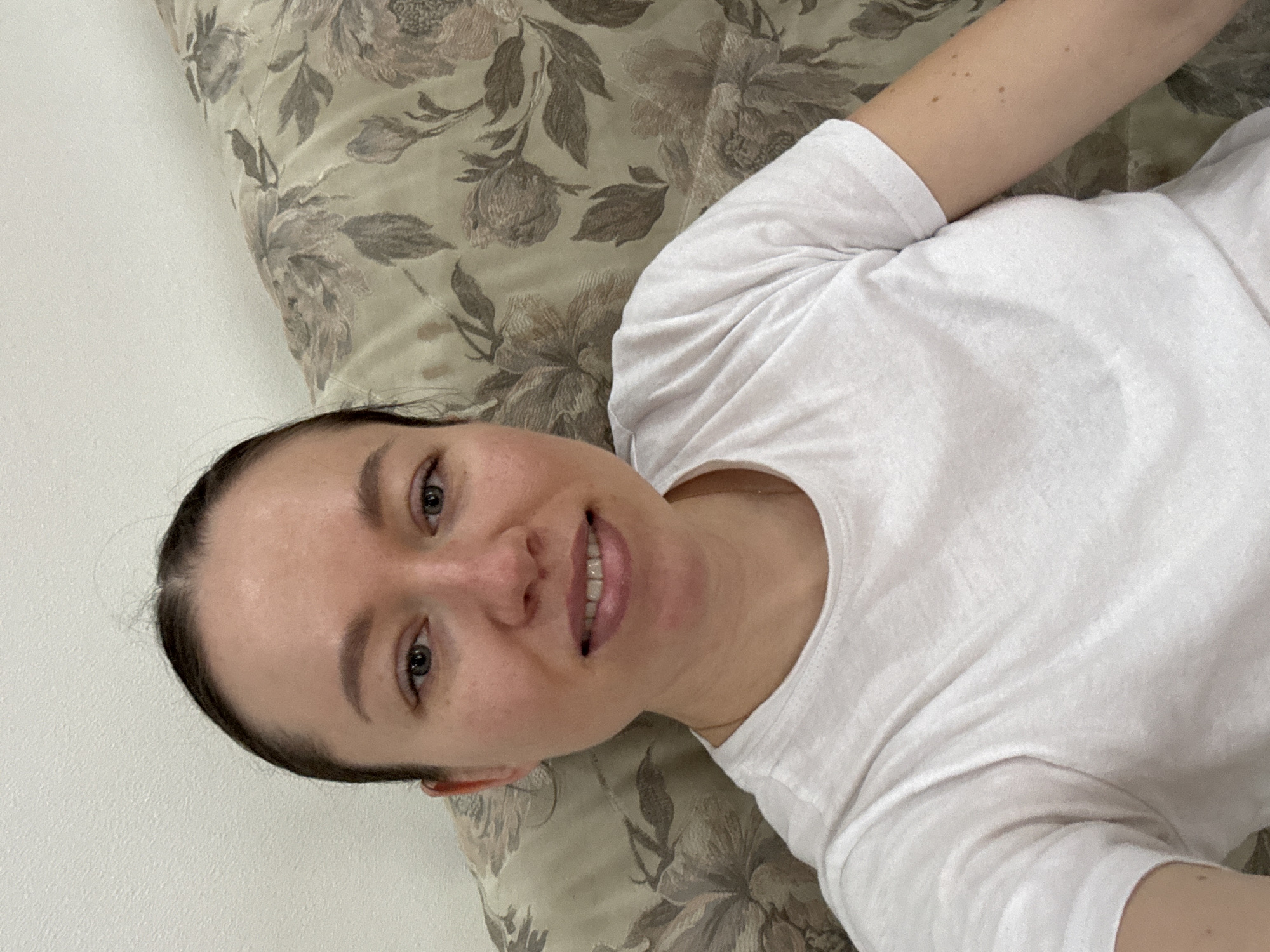 A woman with fair skin, tied-back dark hair, and wearing a white T-shirt is lying on her side on a floral-patterned cushion. She is smiling slightly and looking at the camera. The background includes a plain white wall. The location is unknown and cannot be determined from the image.
