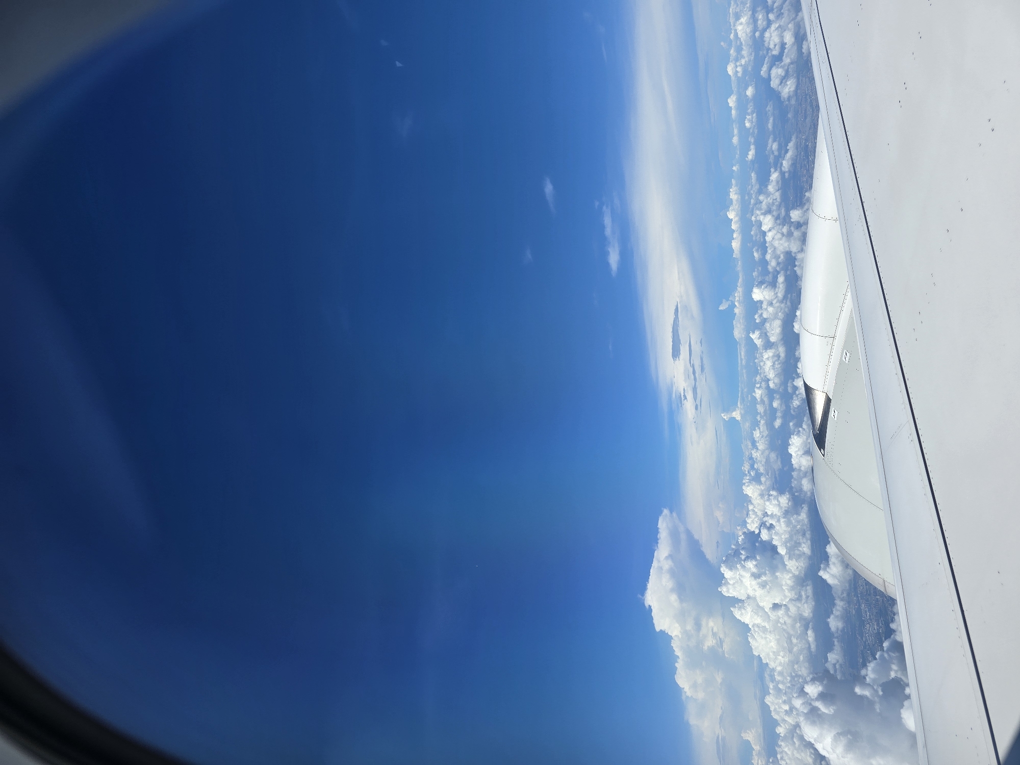 An aerial view from an airplane window shows a vast expanse of bright blue sky with scattered white clouds. In the foreground, a portion of the airplane's wing and engine are visible. Below the clouds, the urban landscape of Dongguan, China, can be faintly discerned, with buildings and city structures appearing small and distant. The scene captures a moment during an airplane journey over the city.