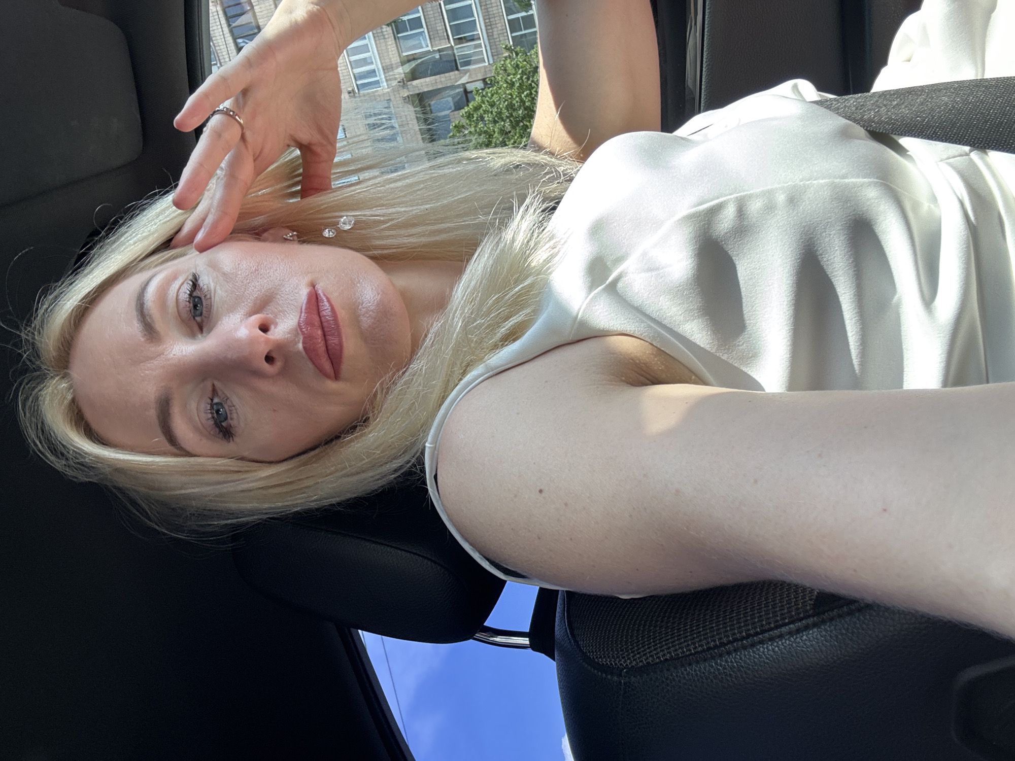 A woman with blonde hair is sitting in the driver's seat of a car, looking at the camera. Her right hand is raised to her forehead, and she is wearing a white dress. The background shows a blue sky, trees, and buildings, suggesting an urban environment. The image is taken during the daytime. The location is Vaganovo, Russia.