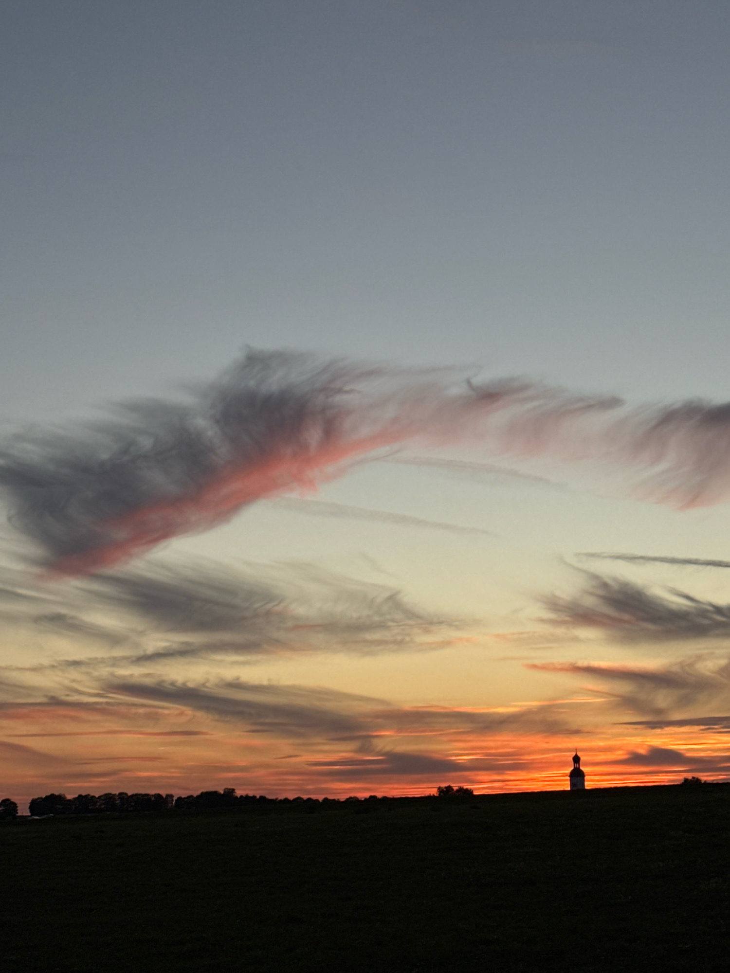 A silhouetted chapel stands on a hilltop against a dramatic sunset sky. The sky is a gradient of light blue at the top, transitioning to warm orange and red hues near the horizon. Wispy clouds are scattered across the sky, with some catching the sunset light and appearing pink and orange. In the foreground, a dark, silhouetted field stretches towards the horizon. The chapel has a distinct dome-shaped roof and appears to be the only man-made structure visible. The overall scene conveys a sense of peace and natural beauty at dusk. The image is likely taken in Wettstetten, Germany, given the presence of a chapel on a hilltop, a common feature in the Bavarian landscape.
