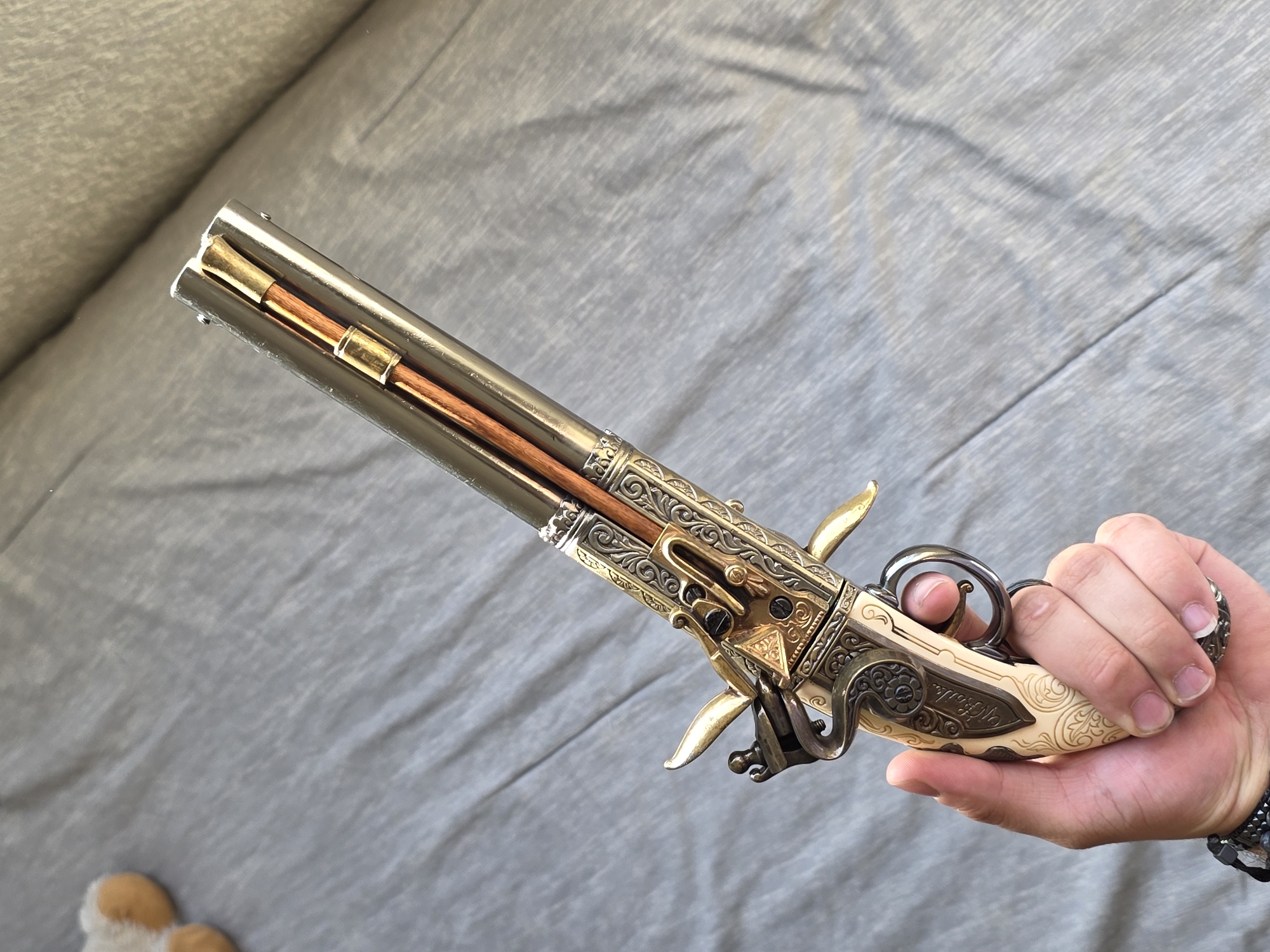 A hand holds an ornate antique pistol, possibly a replica, featuring a metal barrel, engraved gold-colored mechanisms, and an ivory-colored handle with decorative patterns. The hand has pale skin, short nails, and a dark bracelet. The background is a gray, textured surface resembling a bed or couch, and a small portion of a stuffed animal is visible in the lower left corner. The image suggests a display or examination of the firearm in a domestic setting. No specific city or location details are discernible from the visual information.