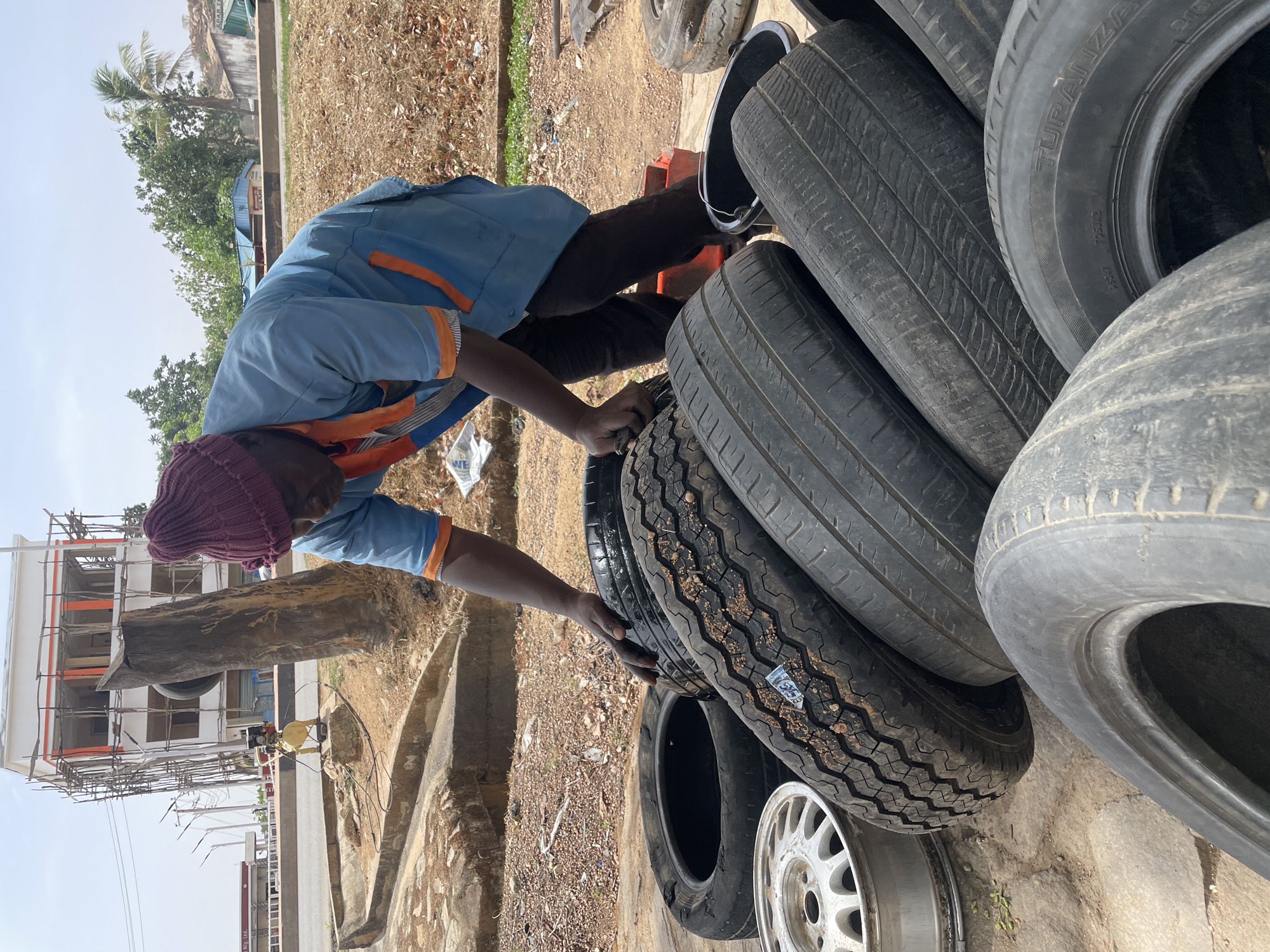 An outdoor scene in Jos, Nigeria, features a man among a stack of used car tires, suggesting a tire repair shop or roadside service. The man, likely a mechanic, is dressed in a light blue long-sleeved shirt with orange accents, dark trousers, and a purple knit beanie. He is actively engaged with the tires, with his hands placed on one, possibly in the process of moving or inspecting it.
Numerous black rubber tires, some fitted with metal rims, are stacked around the man. One tire clearly displays the brand "TURANZA" and tire specifications like "215/60R16" and "TREADWEAR". Another tire has a small sticker with "FALKEN" visible. The ground is a mix of dirt and concrete, with some scattered debris and small green plants. A white bag with partial text "WE" is visible near the man's left foot.
In the background, a multi-story building under construction, featuring scaffolding, is visible. Further back, there are several trees, including a prominent palm tree. A large, dark tree trunk stands upright, partially obscuring the view. A yellow pump or generator-like machine connected by pipes is also present on the ground. The bright, clear daylight indicates a sunny day, likely mid-morning or afternoon. The overall impression is one of a bustling, functional work environment in an urban or semi-urban setting.