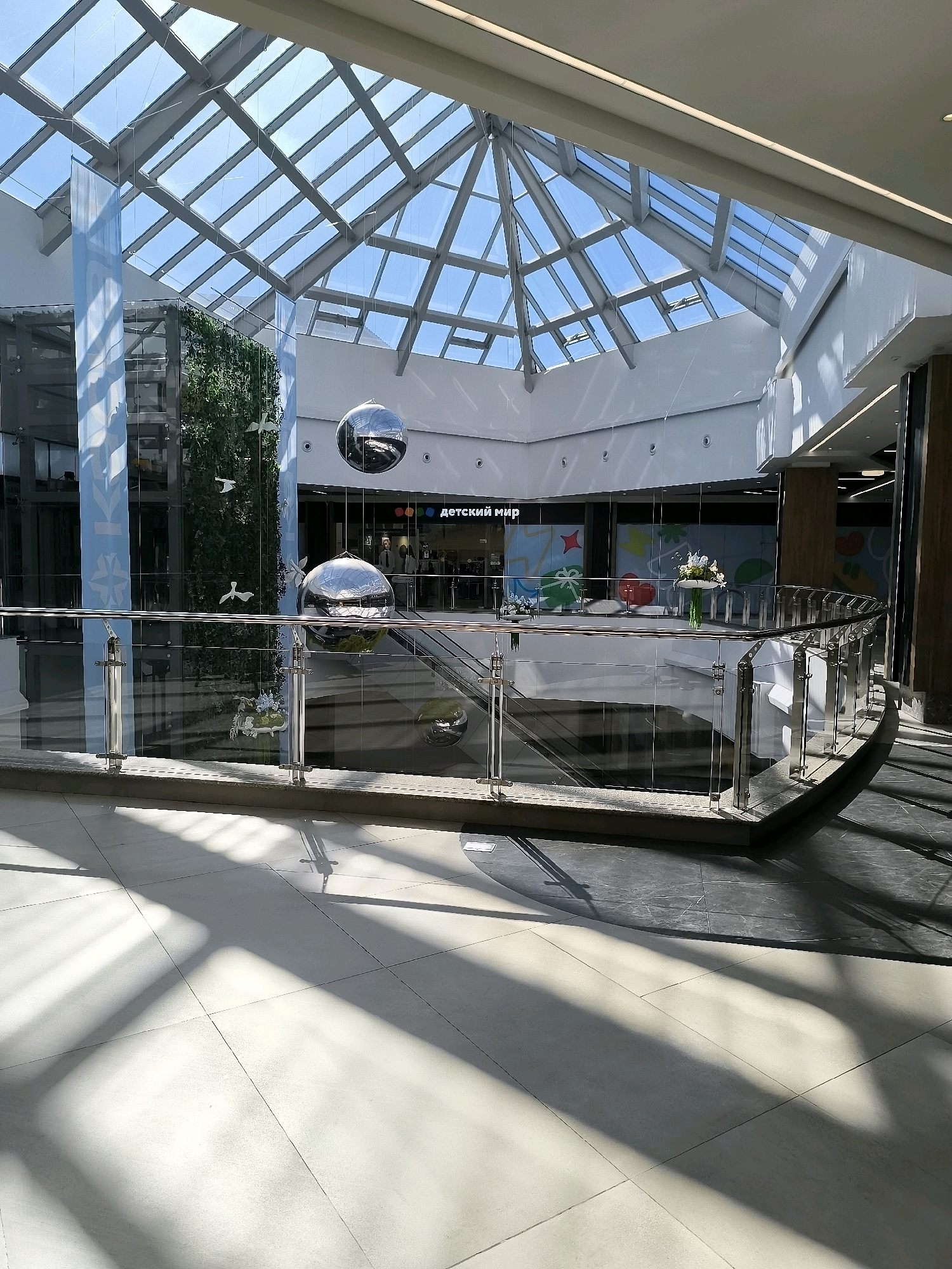 This is an interior shot of a modern shopping mall, characterized by a large, geodesic glass roof that allows abundant natural light to flood the space. The ceiling structure is a complex geometric framework of metal beams supporting translucent panels. Below the roof, large, spherical, mirrored art installations hang suspended, reflecting the surrounding environment.
A vertical green wall, resembling a living moss installation, is visible on the left side of the frame, interspersed with decorative white bird figures. Blue banners with a subtle pattern hang alongside it. In the background, a store with a sign reading "детский мир" (Children's World) is visible. The mall features multiple levels, indicated by a curved railing made of glass and metal. Escalators are also present, suggesting vertical circulation within the mall. The flooring consists of large, light-colored tiles in the foreground, with darker tiled flooring in the mid-ground. Shadows from the overhead structure are cast dramatically across the floor. The scene is clean and well-maintained. There are no people clearly visible in the image. The location is identified as the city of Qarağandı, Kazakhstan.