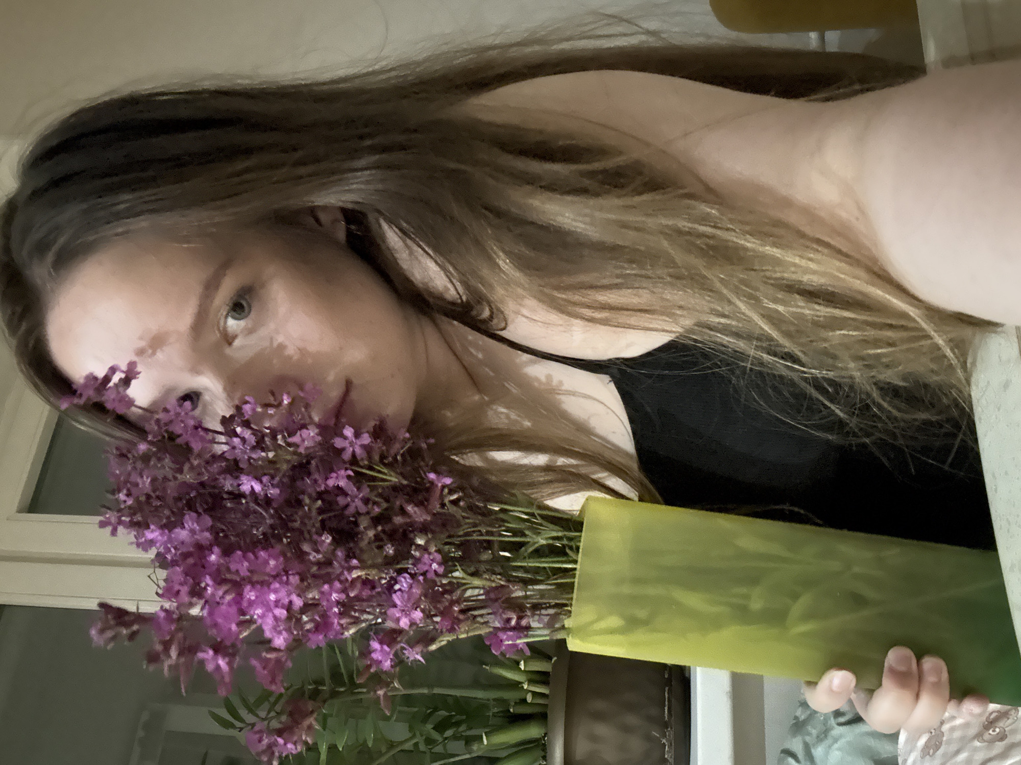 A young woman with long, light brown hair and light-toned skin is positioned centrally in an indoor, close-up shot. Her left eye, which is light-colored, looks directly at the viewer. A dense bouquet of small, vivid purple flowers held in a tall, translucent lime-green vase partially obscures the left side of her face. She wears a black spaghetti-strap top, revealing her right shoulder and upper chest. Her right hand grips the lower part of the vase. In the background to the left, a dark brown potted plant, possibly a Zamioculcas zamiifolia with several cut stems, rests on a surface. A white window or door frame is visible behind her head and the flowers, with a light-colored wall to the right. The foreground shows a light-colored surface beneath the vase and the woman's hand, along with a patterned, light-colored fabric in the bottom left corner. The scene is illuminated by soft, indoor lighting.