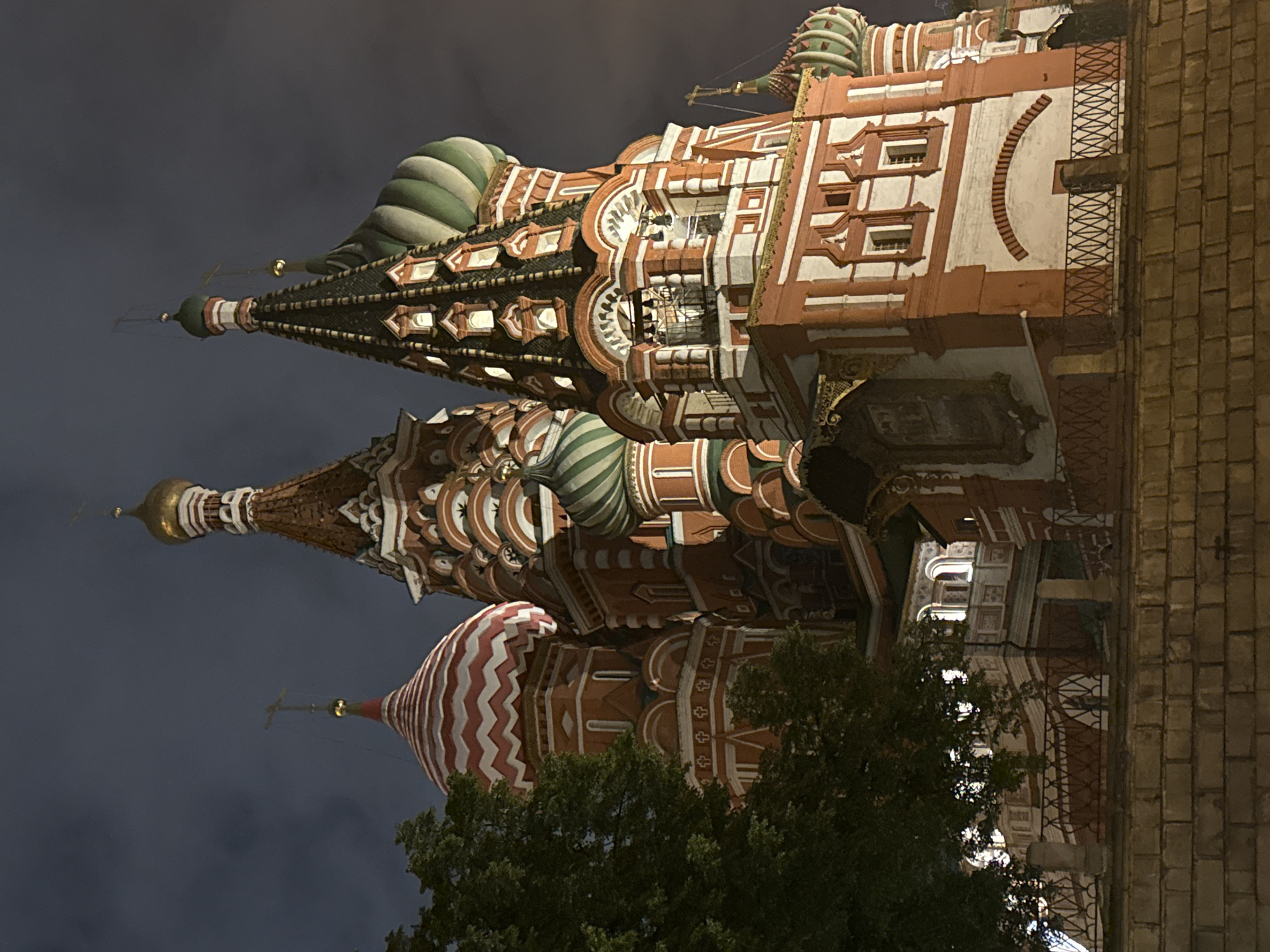 This image shows St. Basil's Cathedral in Moscow, Russia at night. The building's onion domes and ornate exterior are visible, with some trees obscuring the lower portion. The sky is dark, and the cathedral is illuminated, highlighting its architecture. A portion of a brick wall is seen on the right side of the image, suggesting the cathedral's location within the city's urban environment.