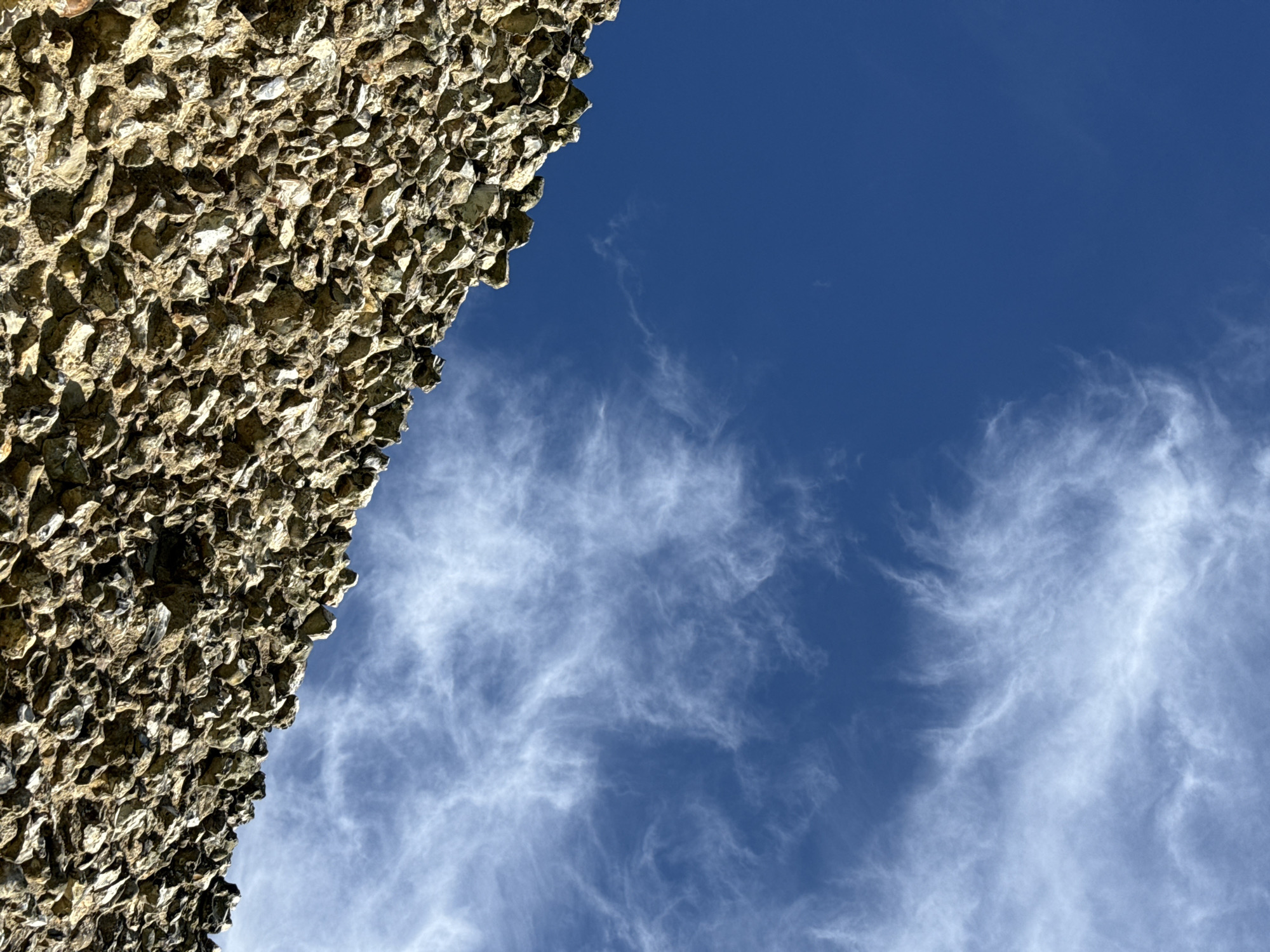 A low-angle shot shows a section of wall constructed of small, rough-hewn, irregularly shaped stones or shells, perhaps flint nodules, on the left side of the frame, contrasting with a bright blue sky with thin, wispy cirrus clouds on the right. The structure appears to be a part of a building. It is located in the city of Dacorum, United Kingdom.