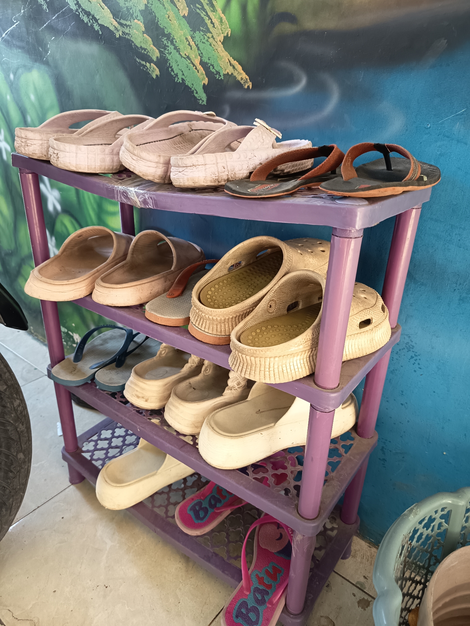 A purple, three-tiered shoe rack is positioned against a blue wall with a green, abstract mural in Gebangan, Indonesia. The rack holds several pairs of footwear. The top shelf displays four pairs of beige slip-on sandals, two of which are slightly offset from the others, and two pairs of brown flip-flops. The middle shelf contains three pairs of beige clogs with textured insoles, one pair of beige slip-on sandals, and one pair of blue and gray flip-flops. The bottom shelf holds a pair of white slip-on sandals and two pairs of pink flip-flops with multicolor designs. To the left of the shoe rack, a portion of a black tire is visible, and to the right, a light blue plastic basket is present. The floor is tiled with a light-colored pattern.