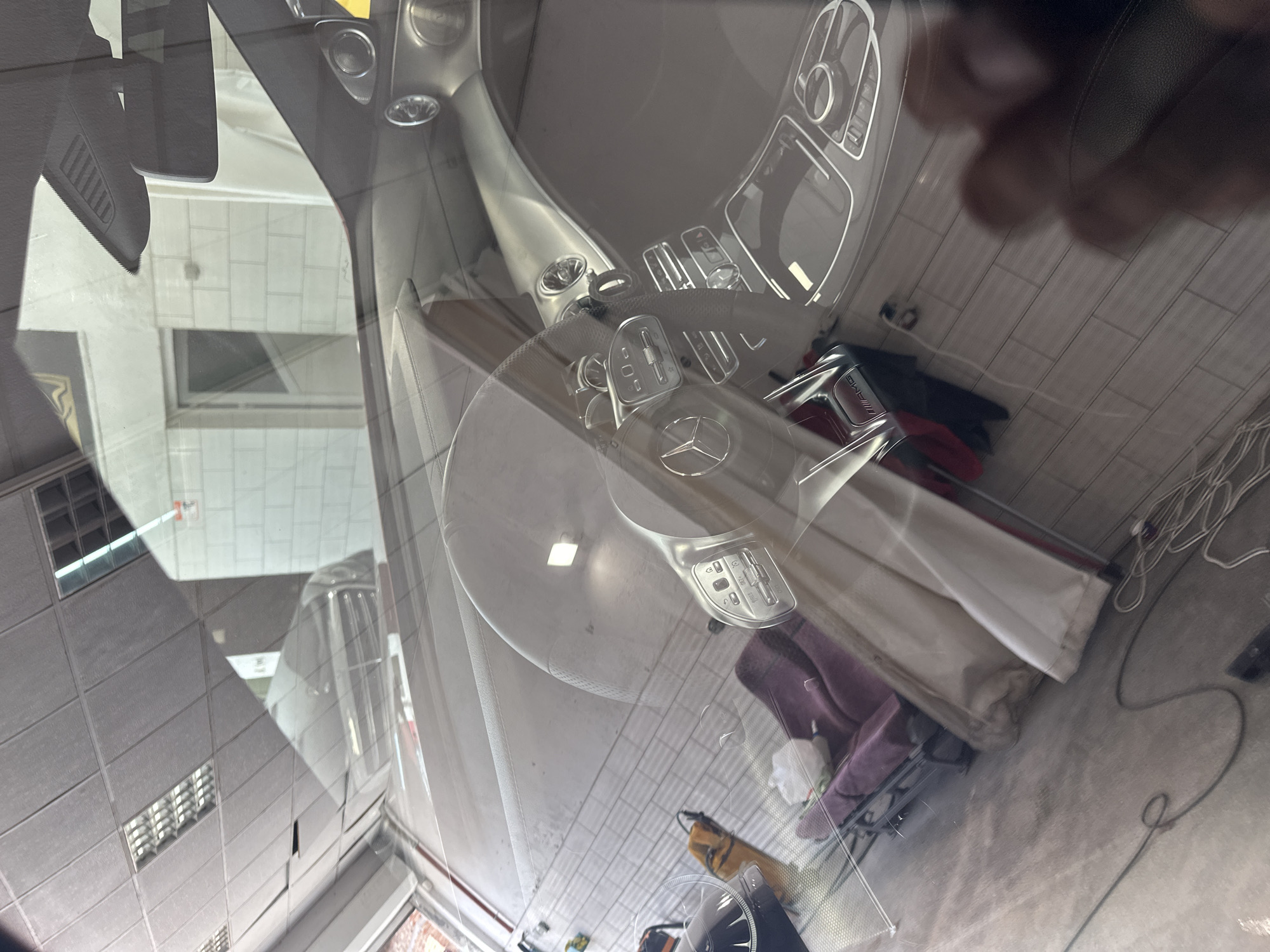 The image shows the interior of a car, viewed through the windshield. The car appears to be parked indoors, possibly in a garage or workshop, with a concrete floor and some lighting visible in the background. The interior features a sleek, modern dashboard with a prominent Mercedes-Benz logo on the steering wheel. There are also various control buttons and panels visible. A portion of a person's hand is visible at the top right of the image, suggesting someone is holding the phone used to take the picture. The overall impression is one of luxury and high-tech automotive design. The lighting suggests it might be daytime, but the indoor setting makes it difficult to determine the exact time or weather. No text is clearly legible within the image.