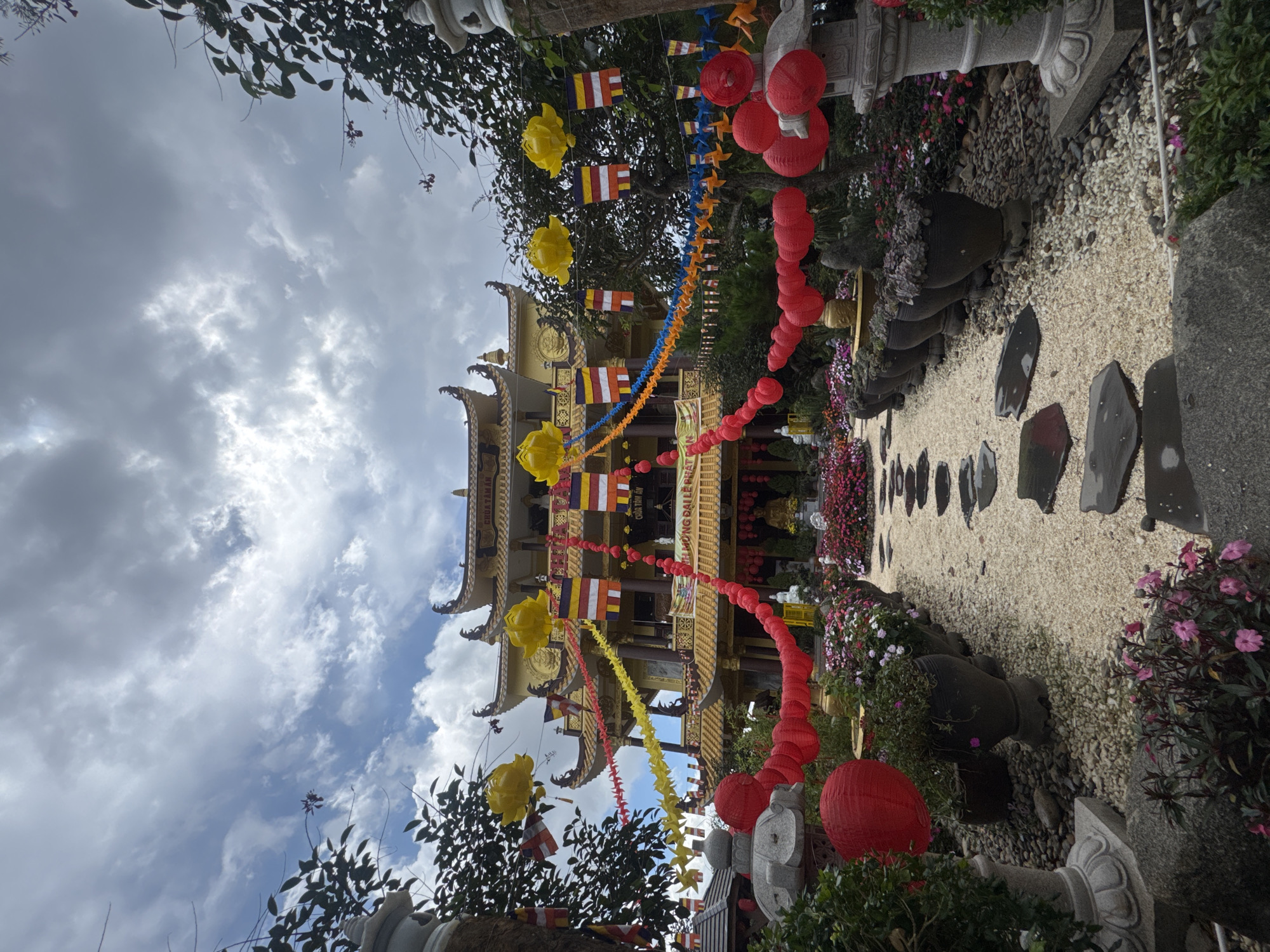 The image depicts a vibrant Buddhist temple, identified as Chua Tam An, located in Da Lat, Vietnam. The temple is a multi-tiered structure with yellow tiled roofs and ornate architectural details. It is adorned with colorful decorations, including yellow lotus lanterns, Buddhist prayer flags, and strings of red paper lanterns. A pathway paved with light-colored gravel and accented with flat, dark stepping stones leads towards the temple entrance. The path is flanked by lush greenery, potted plants, and an abundance of blooming flowers in shades of pink and white. Several large stone urns and decorative stone lanterns are visible along the path and in the garden areas. The sky above is partly cloudy with patches of blue sky and dramatic grey clouds. The overall scene conveys a sense of tranquility and spiritual ambiance, characteristic of a place of worship and contemplation. There are no people visible in the image.