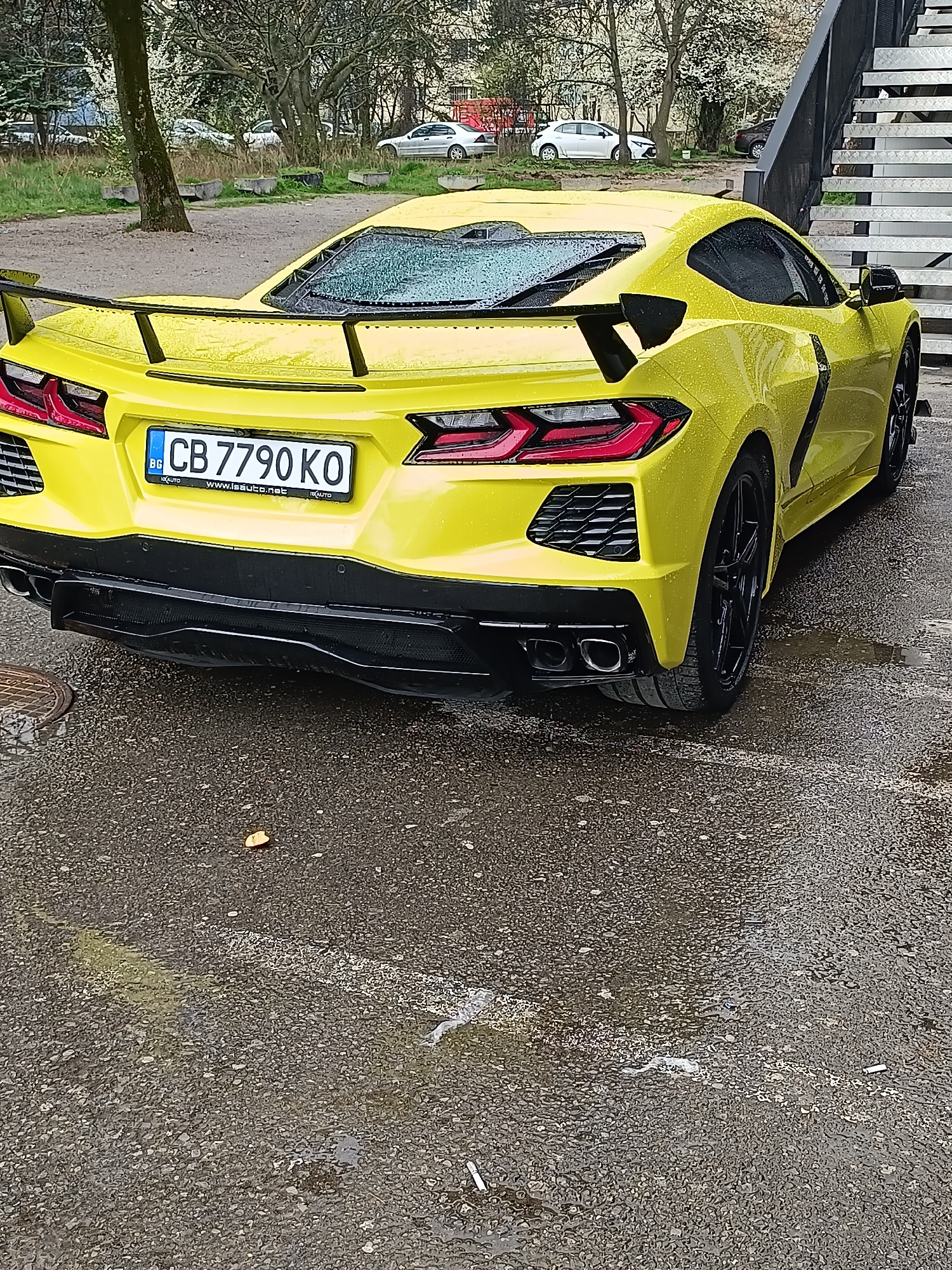 A yellow sports car, likely a Chevrolet Corvette C8, is parked on a wet asphalt surface. The car has a prominent black rear wing and black wheels. The license plate reads "CB 7790 KO" and is of Bulgarian origin. The background includes trees and some buildings, suggesting an urban or suburban setting. The stairs of an unidentified building are visible on the right. Based on the license plate, the location is within Bulgaria, and judging from the modern building elements, it could be in Sofia.