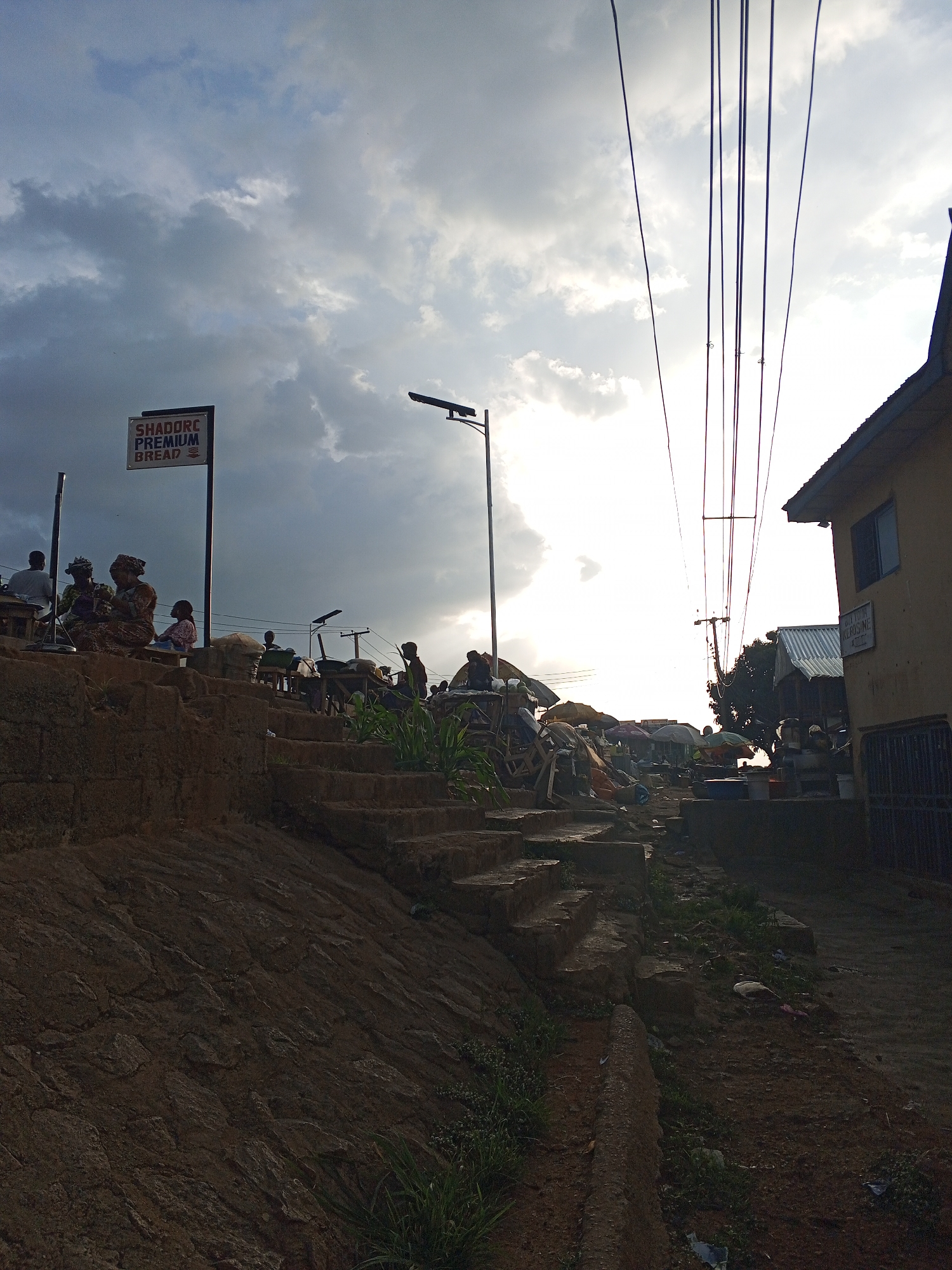 The image shows a street scene in Gwong, Nigeria. A weathered stone wall forms the left foreground, leading up to a set of concrete steps. People are sitting and standing near a sign that reads "SHADORE PREMIUM BREAD." The street is lined with stalls and vendors under umbrellas, indicating a market. Power lines stretch across the sky. A building is visible on the right side of the street. The sky is cloudy with the sun's light peaking through.
