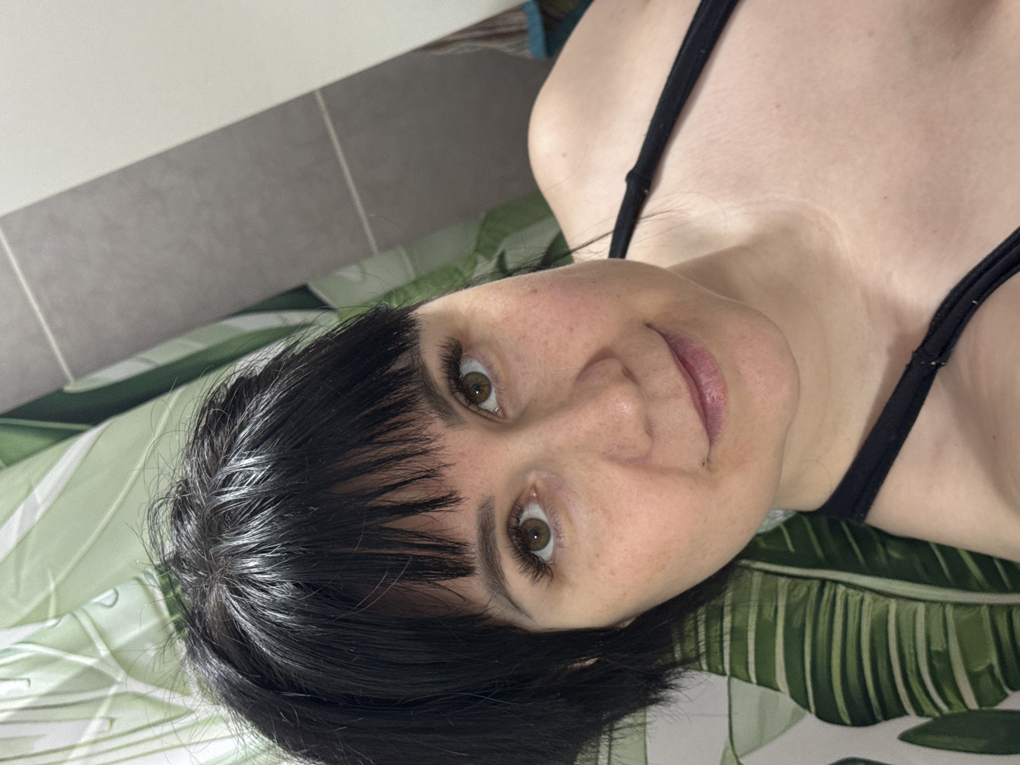A person with dark hair and brown eyes is lying on a surface with a green and white botanical print. They are wearing a black tank top. The background includes a section of grey tiles and a white surface. There is no information in the image to determine the location in the city of Zav'yalovo, Russia.