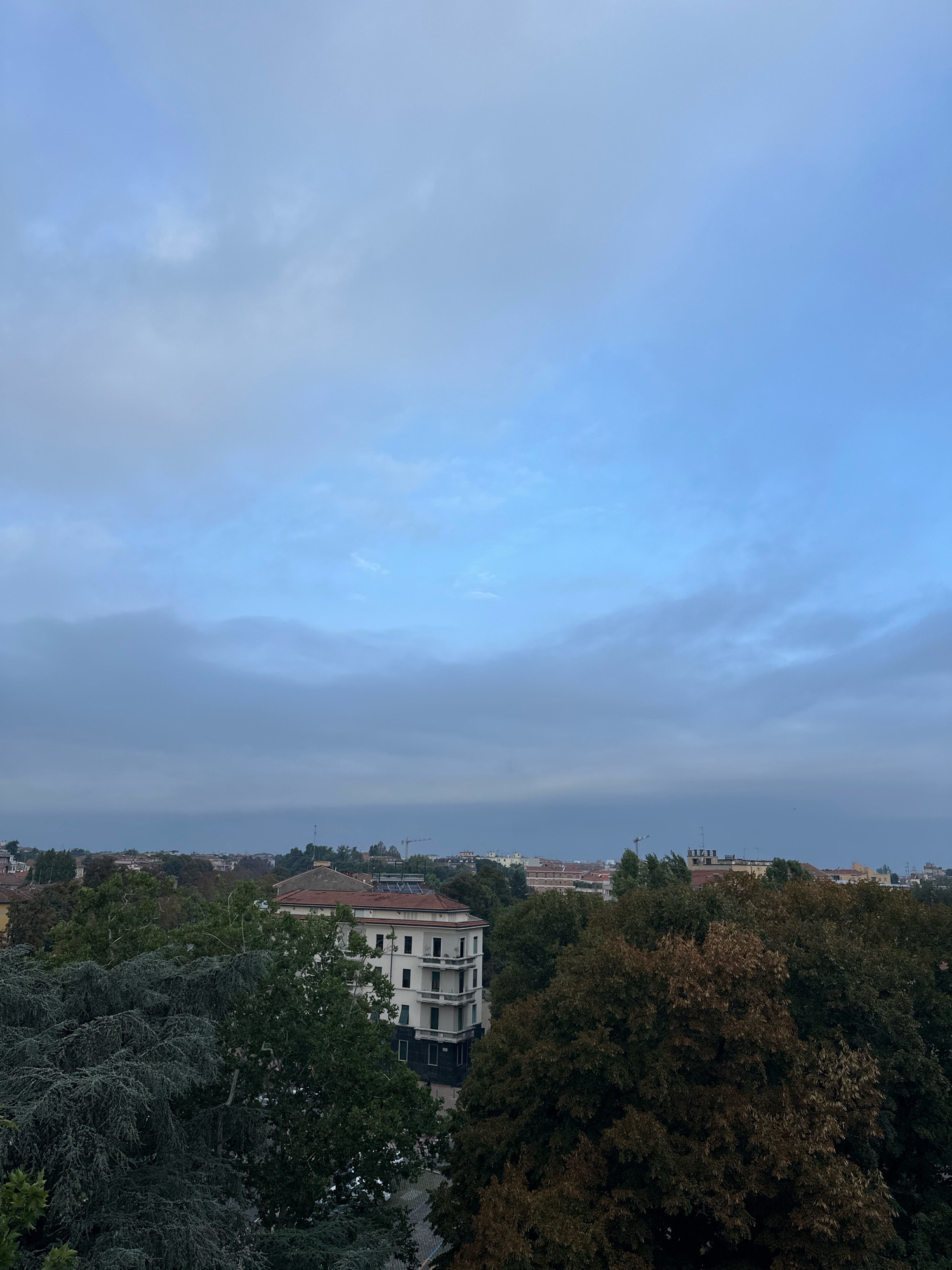 The image shows an aerial view of a city, likely Parma, Italy, characterized by a mix of buildings and lush greenery. The sky is overcast with light blue and grey clouds, suggesting it could be early morning or late afternoon. In the foreground, a large building with a white facade and balconies stands out, partially obscured by dense trees. The trees are a mix of evergreen and deciduous, with some showing autumnal colors. Beyond these, a cityscape is visible with terracotta-tiled roofs and other structures. In the distance, a construction crane indicates ongoing development. The overall scene is tranquil, with no visible people or animals.