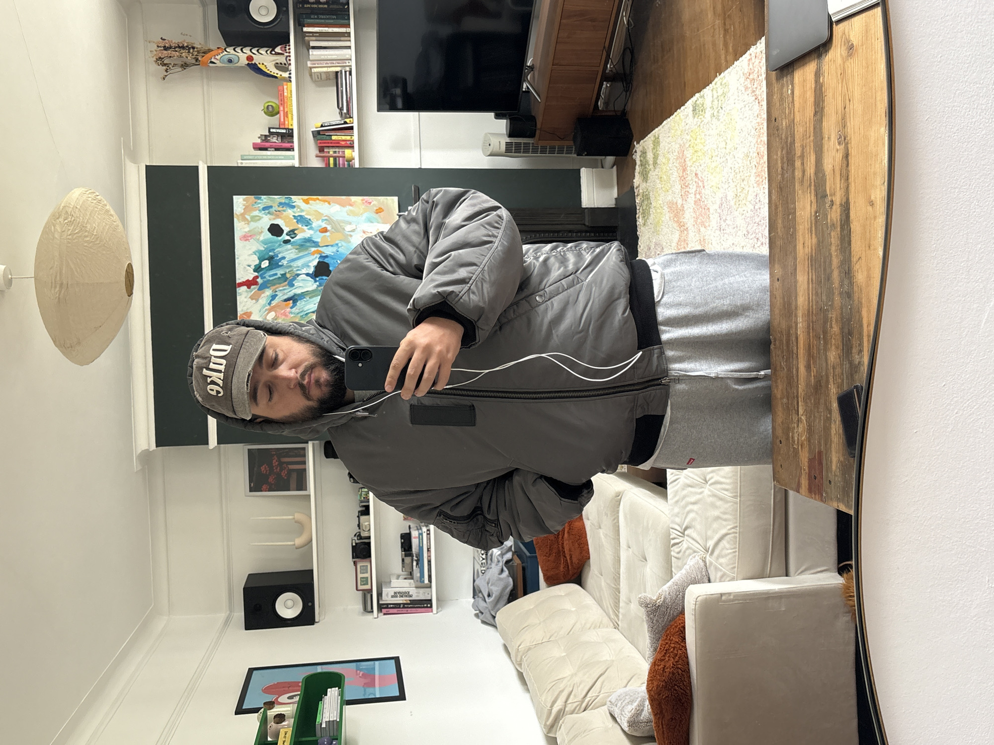A man stands in what appears to be a London residence, holding a smartphone. He is reflected in a mirror, capturing his image and the environment behind him. He is wearing a grey jacket and sweatpants, with a cap on his head. The room features a sofa, a bookshelf filled with books and decorative items, a television, and a painting on the wall. A lampshade hangs from the ceiling. The presence of a fireplace suggests that the scene takes place in a residential living room, possibly in London, given the style of interior design common in UK residences.