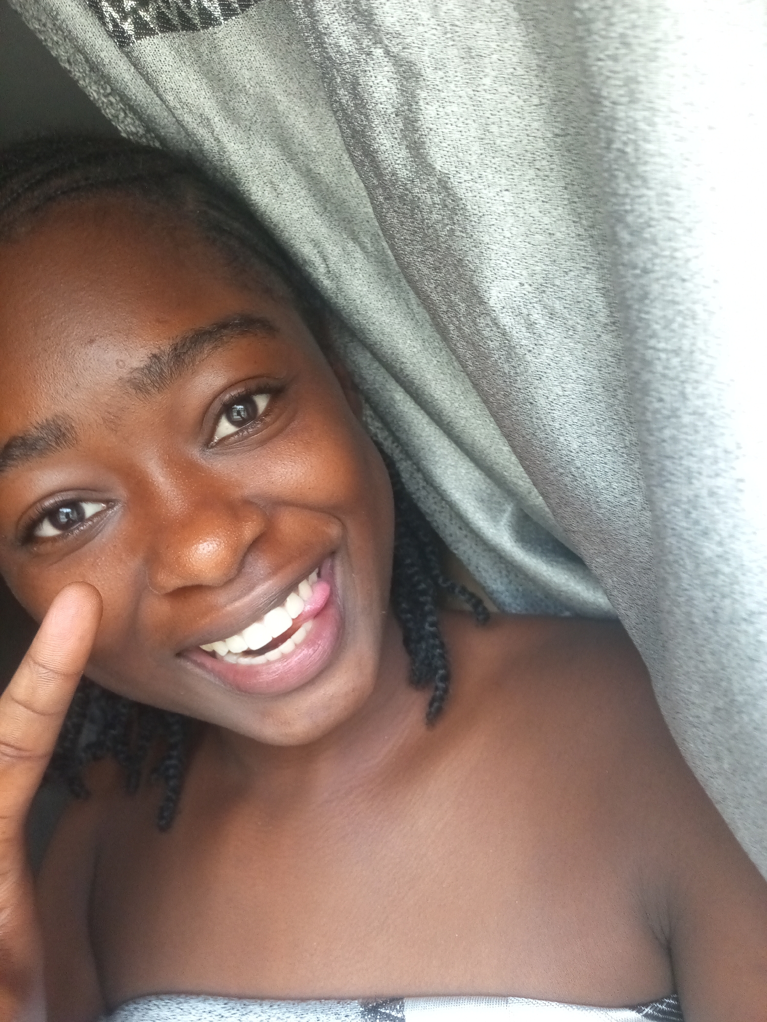 In a close-up, waist-up shot, a young Black woman with dark skin and braided hair smiles with her tongue playfully sticking out. She is positioned in the lower right quadrant of the frame, with her head tilted and her eyes looking directly at the camera. Her mouth is open, revealing bright white teeth and a pink tongue. A thumb is held up near her left cheek, partially obscuring her face. She is bare-shouldered, wearing what appears to be a patterned fabric draped around her upper chest. The background is dominated by a large, textured gray curtain, which creates a sense of enclosure and privacy. The lighting is soft, highlighting the sheen on her skin and the texture of the curtain.