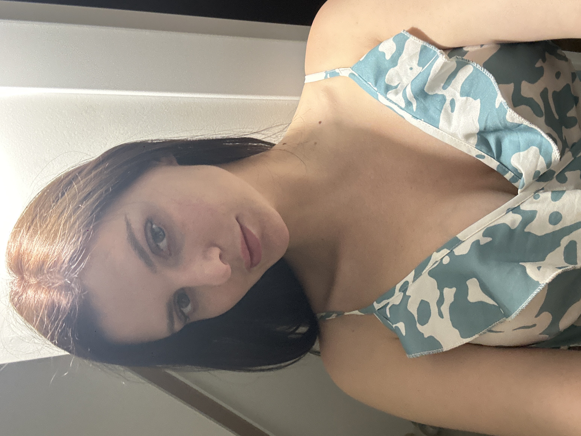 A medium shot shows a young woman with dark brown hair and blue eyes, looking directly at the camera. She has a light complexion and is wearing a light blue and white patterned dress. The background is a plain, light-colored wall.