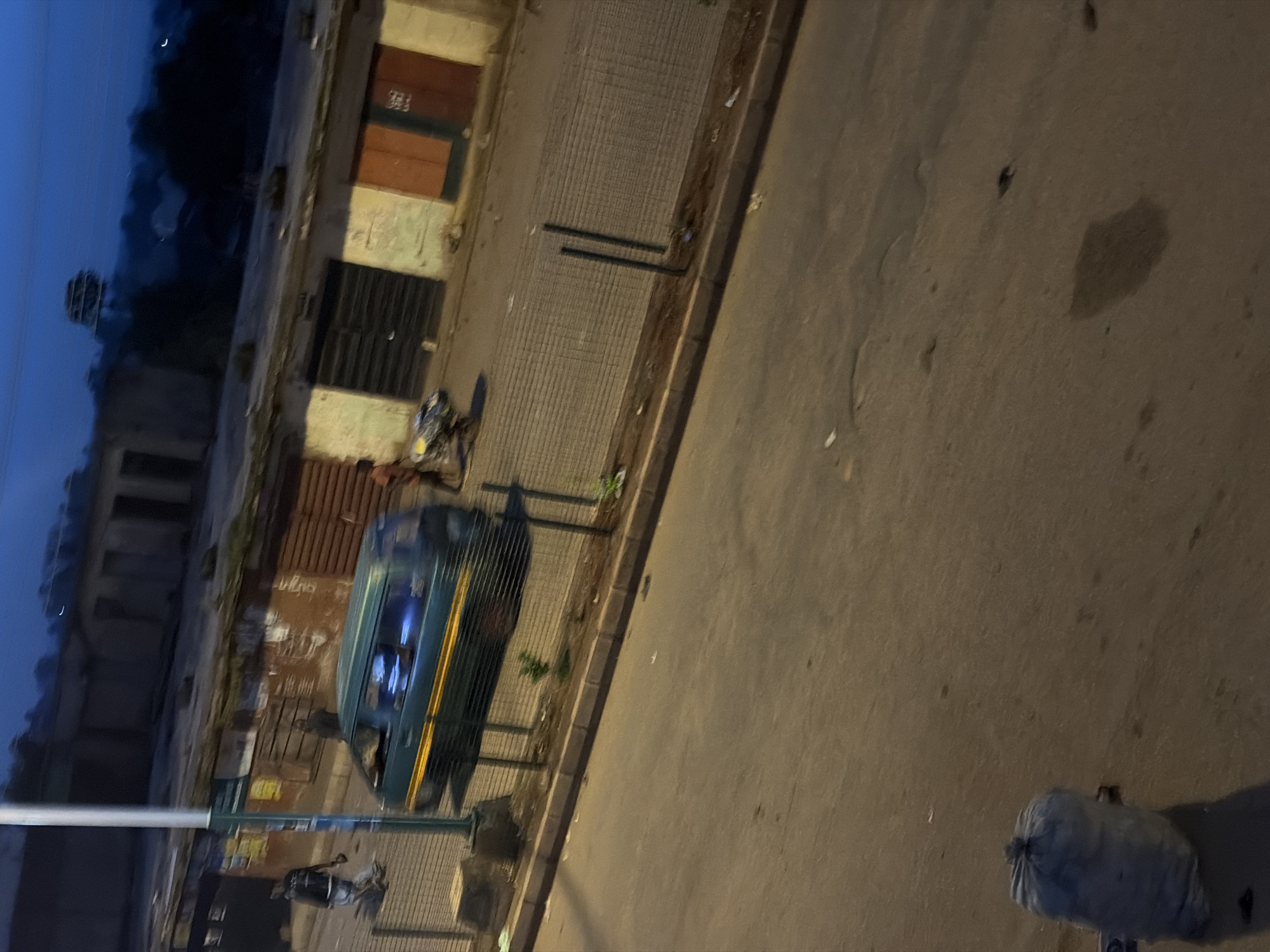 The image depicts an outdoor scene at dusk, likely in Bukuru, Nigeria. The setting appears to be a street or walkway adjacent to buildings. In the foreground, a blue and green vehicle is parked. Further down the street, a person is visible near the vehicle. To the right of the street, a large, light-colored bag, possibly filled with trash, rests on the ground. The sky is dark with a sliver of a crescent moon visible, indicating it is either early morning or evening. The overall lighting suggests a low-light condition. The ground is paved with what looks like cobblestones or rough concrete near the buildings, transitioning to asphalt on the road. The buildings have a worn appearance with some doors and windows visible. There is no discernible text in the image. The image is tilted, with the ground appearing to run diagonally from the bottom right to the top left.