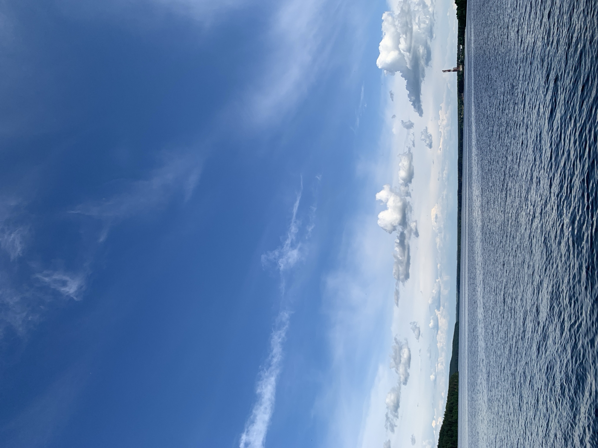 This media file captures a wide panoramic view of a vast body of water under a bright blue sky, oriented horizontally. The scene appears to be taken from a relatively low vantage point, possibly from a boat or the water's edge.
The upper portion of the image is dominated by a clear, azure sky, transitioning from a deeper blue at the top to a lighter hue towards the horizon. Scattered clouds populate the sky; some are high, wispy cirrus clouds, while others are more defined, fluffy cumulus clouds clustered along the middle and right side of the frame. Small dark specks, possibly birds, are visible against the blue sky in several areas.
A distant horizon line separates the sky from the water. Along this horizon, a narrow strip of dark green land, heavily forested, stretches across the frame. Towards the right side, rising above the distant land, stands a tall, slender industrial structure, possibly a chimney or tower, featuring a reddish-brown band near its apex.
The lower portion of the image is filled by the expansive water surface. The water is a deep blue-grey, textured with numerous small ripples and waves, suggesting a gentle breeze or current. The light reflects off these ripples, creating varying shades across the surface.
The time of day appears to be bright daylight, likely midday or early afternoon, indicated by the clear lighting and vivid blue sky. The weather is fair, with a mix of clear sky and scattered clouds. No people or visible text are present. The overall atmosphere is one of calm, natural expanse. Given the location context of Chaikovsky, Russia, this is likely a view of the Kama Reservoir or a large river in the region.