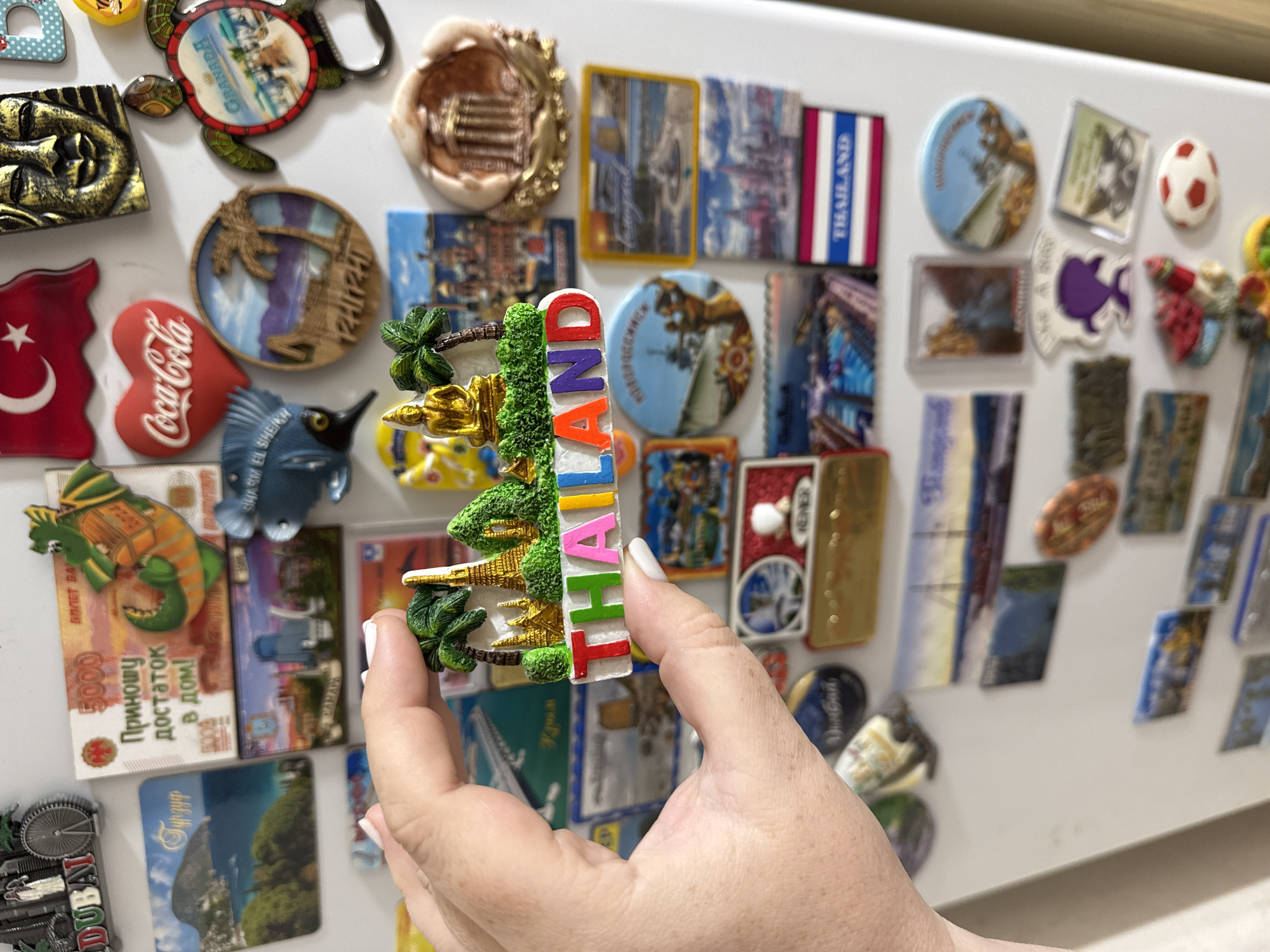 The image is a close-up, top-down view of a person's hand holding a refrigerator magnet shaped like a Thai temple complex with palm trees and the word "THAILAND" in colorful letters. The magnet is positioned in the center of the frame. The background is a white refrigerator door covered with numerous other magnets. These magnets depict various scenes and objects, including landscapes, landmarks, a Coca-Cola logo, a Turkish flag, and a golden Buddha face. The lighting is bright, suggesting an indoor environment. The specific location in the city of Mikhaylovsk, Russia, cannot be determined from the image itself, as the magnets are souvenirs from different locations.