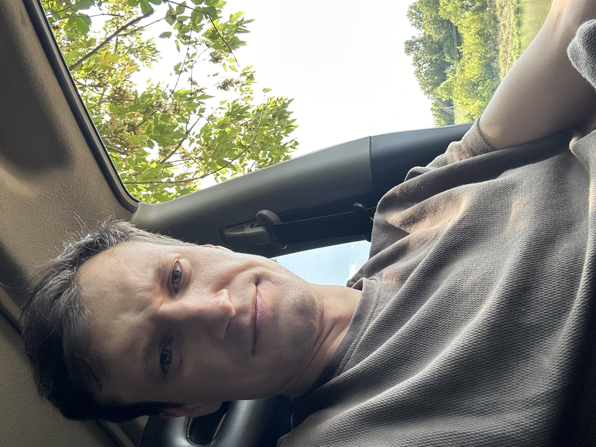 A middle-aged man with short, dark hair is sitting in the driver's seat of a vehicle. He is looking directly at the camera with a neutral expression. He is wearing a brown, textured long-sleeved shirt. The interior of the car is visible, including the black headrest of the seat and a gray pillar on the right side. Through the open window on the right, lush green trees and a body of water are visible under a bright sky. The scene appears to be in a rural setting outside of Makiv, Ukraine.
