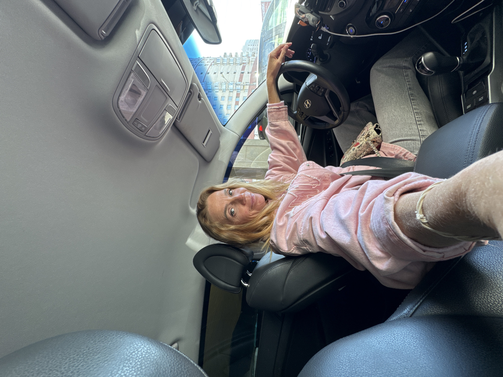 A woman with blonde hair, wearing a pink jacket and grey jeans, lies across the driver's seat of a car. She is wearing a seatbelt. The car interior is visible, including the steering wheel, dashboard, gear stick, and overhead light panel. Outside the car window, a building is visible. Based on the architecture, the location is likely Saint Petersburg, Russia.