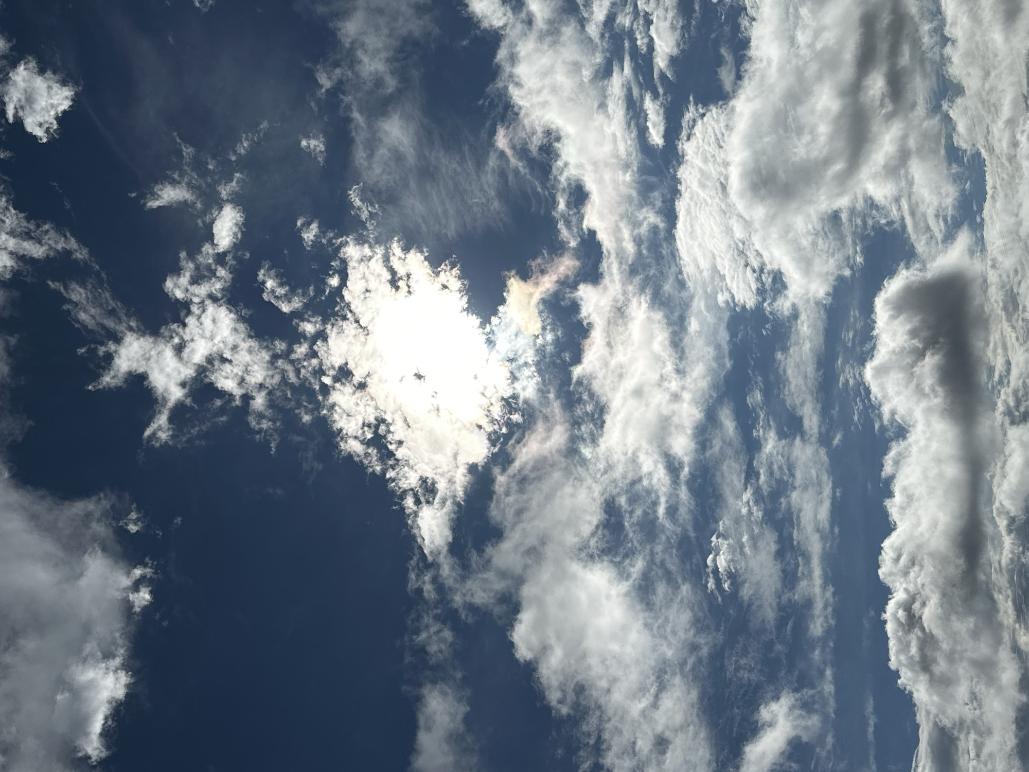 This is a photograph of a partially cloudy blue sky, taken from an outdoor location. The sun is visible through a break in the clouds, creating a bright, hazy area in the upper-middle portion of the image. Wispy, white clouds are scattered across the sky, with larger, more textured clouds present near the bottom of the frame. The image does not contain any specific landmarks, people, or objects that would indicate its location within Podolsk, Russia.
