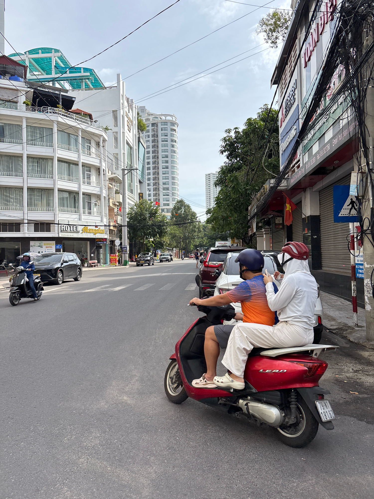 This is a medium shot, eye-level, outdoor, daytime photograph taken in Nha Trang, Vietnam. The foreground features two individuals riding a red Honda Lead scooter. The person in front, wearing a blue helmet and an orange and blue patterned shirt, is driving. The person behind, wearing a red helmet and a white hooded garment with matching white pants, is holding the driver's shoulder. Both are wearing casual footwear. The scooter is on a paved road with a visible license plate. In the background, a line of vehicles, including cars and other scooters, are visible on the road, suggesting traffic. To the right, there is a sidewalk with a pole displaying a pedestrian crossing sign and a Vietnamese flag. Buildings line the street, with a mix of modern high-rises and older establishments featuring signage and balconies. The sky is overcast with some visible clouds. The scene depicts a typical street view of urban transportation and architecture in Vietnam.