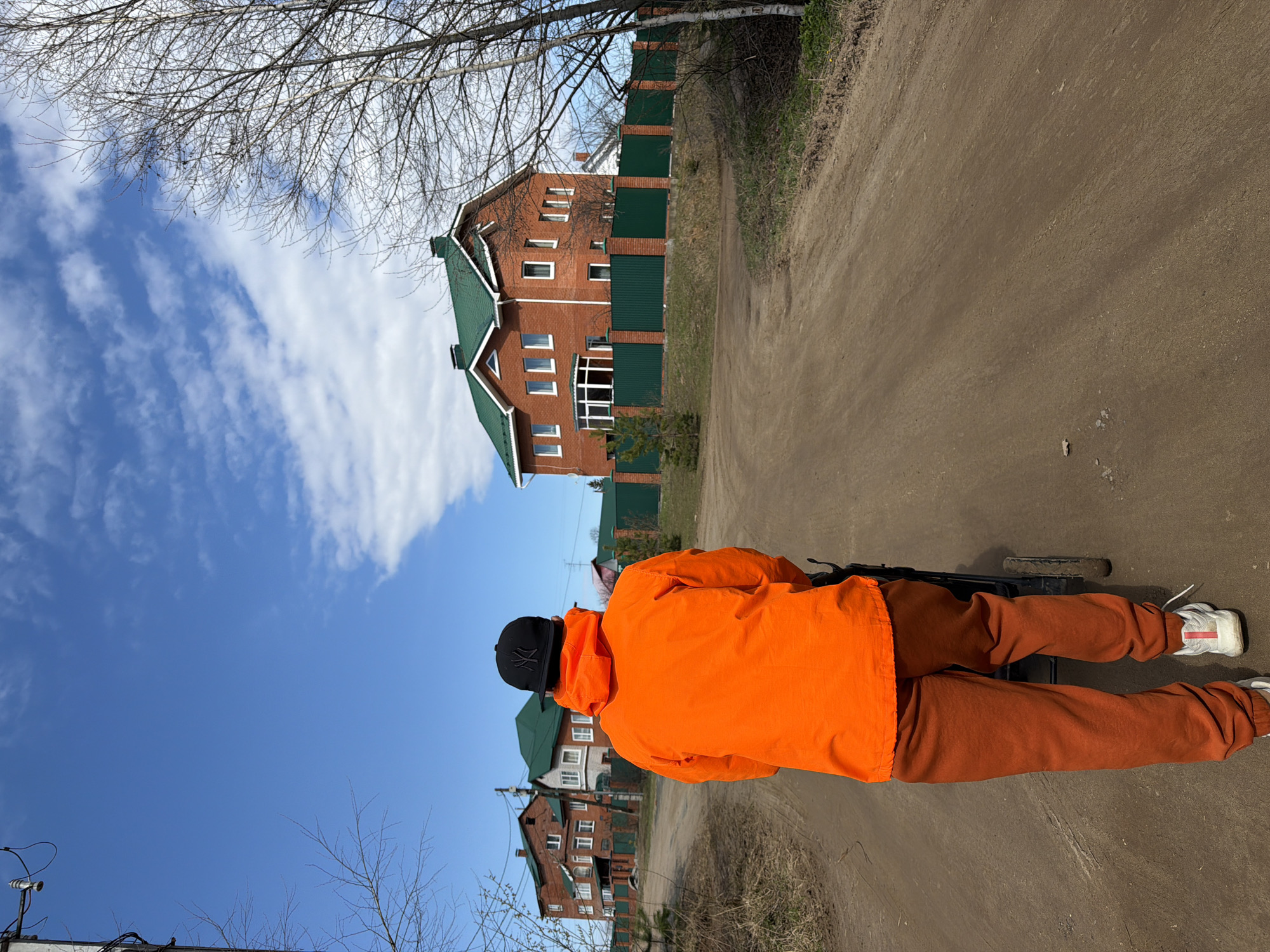 An outdoor, full-length, back view shows a person walking on an unpaved dirt road in Chaikovsky, Russia. The individual wears an orange hooded jacket, matching orange/brown sweatpants, and white sneakers with a red stripe. A black baseball cap with a white "NY" style logo is worn backwards. The person appears to be pushing a dark-colored object, possibly a stroller or cart, partially visible at their feet.
In the background, on the left side of the road, stands a large, multi-story house made of red brick with a dark green gabled roof and white window frames. A tall, dark green fence surrounds the property. Another house with a similar architectural style is partially visible further down the road. The setting suggests a suburban or rural residential area.
Above, the sky is a bright blue with scattered white clouds, indicating clear weather. Bare tree branches, typical of late winter or early spring, frame the upper part of the image. The ground is a light brown dirt road with some sparse vegetation along its edges. The scene captures a peaceful daytime walk under a bright sky.