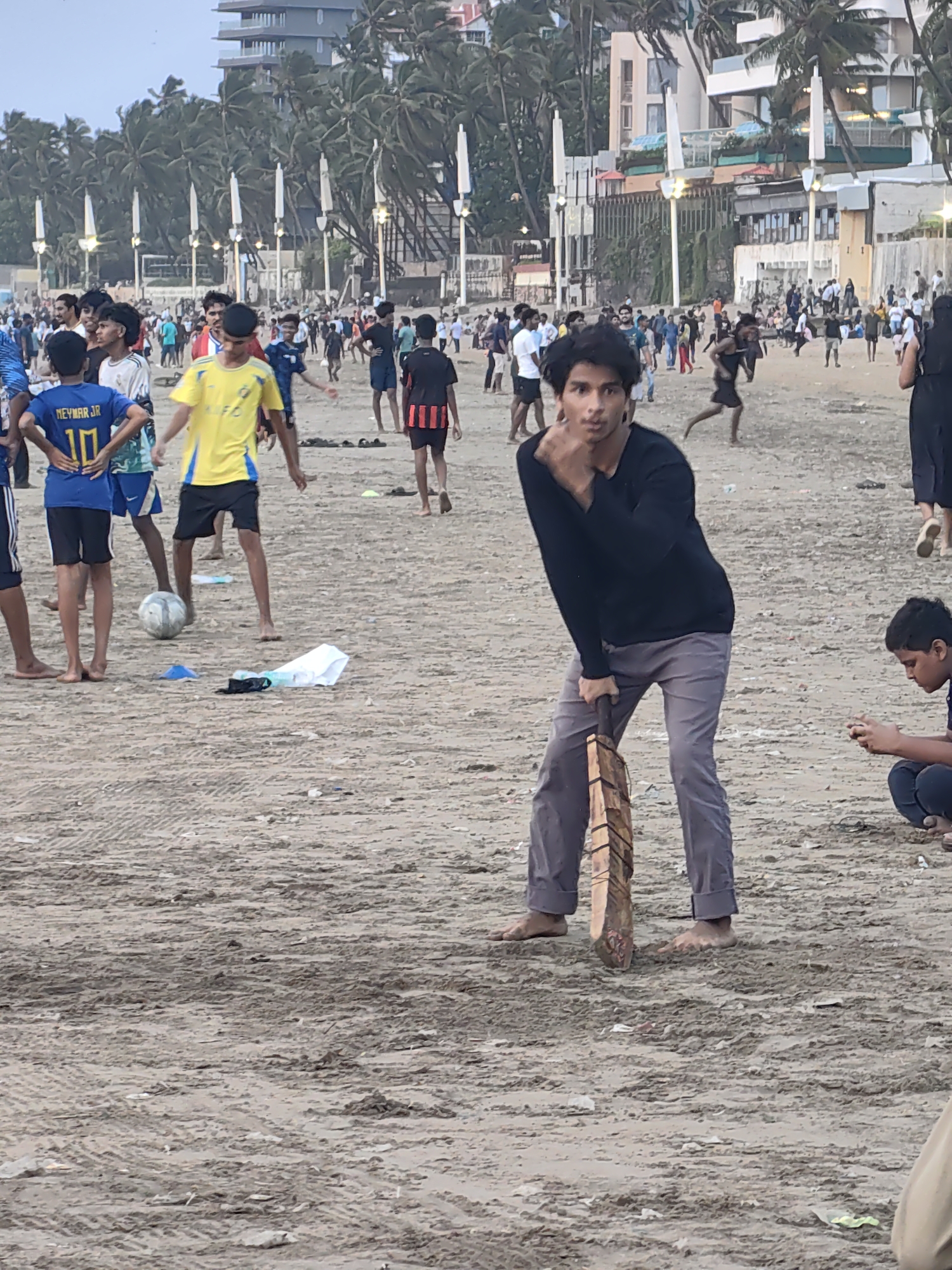 This image captures a lively scene on Juhu Beach in Mumbai, India. In the foreground, a young man in a black long-sleeved shirt and grey pants is holding a cricket bat, seemingly about to play. His posture suggests a moment of anticipation or playfulness. To his right, another young boy is engrossed in his mobile phone.
Further back, a group of individuals are engaged in a game of football, with a soccer ball visible on the sand. There are several other people scattered across the beach, some walking, some standing, and some observing. The background reveals the iconic Juhu Beach skyline, featuring palm trees, buildings, and what appear to be beachfront establishments. The sand itself is textured with tire tracks and strewn with minor debris, indicating a well-used public space. The overall atmosphere is casual and recreational, typical of a beach scene in a bustling city.