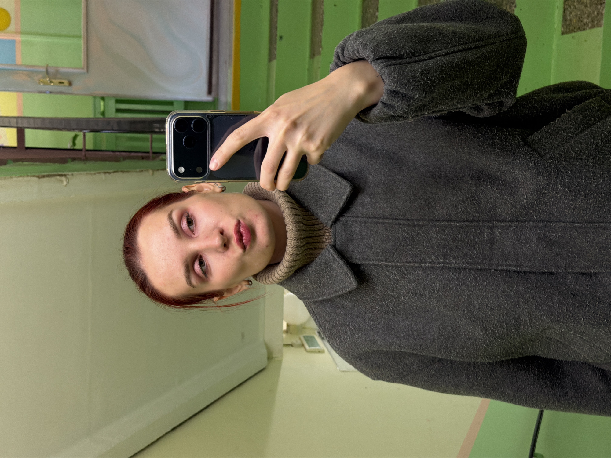 A portrait-oriented selfie captures a young woman looking directly into the camera. She holds a dark smartphone, likely an iPhone due to its distinctive triple camera array, in her right hand, positioned to take the photo. Her left hand is also visible, resting near the phone. She wears a warm, dark grey coat with a noticeably fuzzy or textured fabric, and a light brown turtleneck sweater underneath. Her hair appears reddish-brown, pulled back, and she has visible eye makeup, possibly pinkish eyeshadow, along with a neutral expression and slightly parted lips.
The background suggests an indoor setting, likely a stairwell or hallway in a building located in Chaikovsky, Russia. The walls are painted a light, somewhat muted green, with sections of darker green and hints of other pastel colors, possibly from abstract art or decorative elements on the walls. A dark metal railing is partially visible behind her. The floor or lower wall section is also a light green or beige. The overall lighting appears to be artificial or diffused natural light from indoors. No visible text is present in the image.