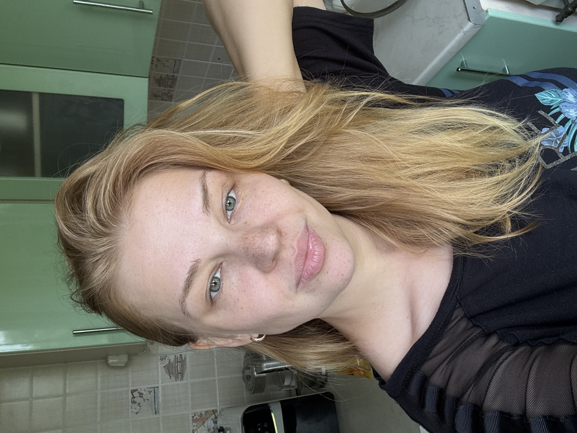 A young woman with blonde hair and fair skin is taking a selfie in a kitchen. She is wearing a black t-shirt with sheer sleeves. Her hair is loose and falls over her shoulders. She has green eyes and a smattering of freckles across her nose and cheeks. Her lips are slightly parted, and she is looking directly at the camera with a neutral expression. The background shows kitchen cabinets, tiled walls, and some appliances. The lighting suggests it is daytime. The setting is a domestic interior, likely a kitchen, with standard cabinetry and wall tiles.