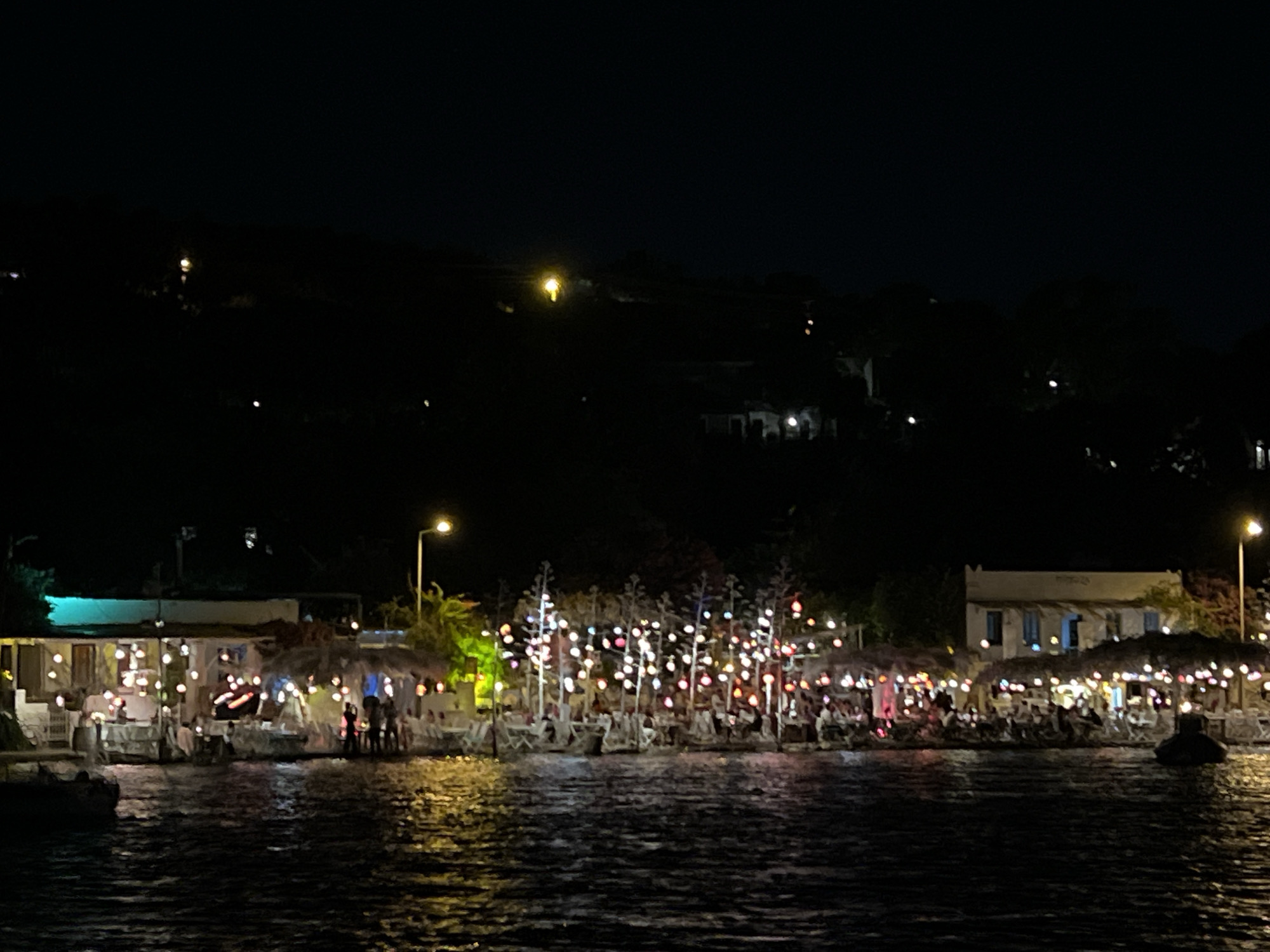A nighttime, wide shot captures a waterfront scene at an unknown location. A body of water, likely a sea or large lake, fills the foreground, reflecting the lights from the shore. The shore itself is lined with several low-rise buildings and structures, possibly restaurants or cafes, adorned with numerous colorful string lights. These lights create a festive atmosphere, illuminating thatched roofs and outdoor seating areas where numerous patrons are visible, though their features are indistinct due to the low light. In the background, a dark, undulating hillside is dotted with distant lights, suggesting residential areas or other establishments. A single, bright streetlight stands to the right of the frame, casting a pale glow. The overall impression is one of a lively, illuminated waterfront destination at night.