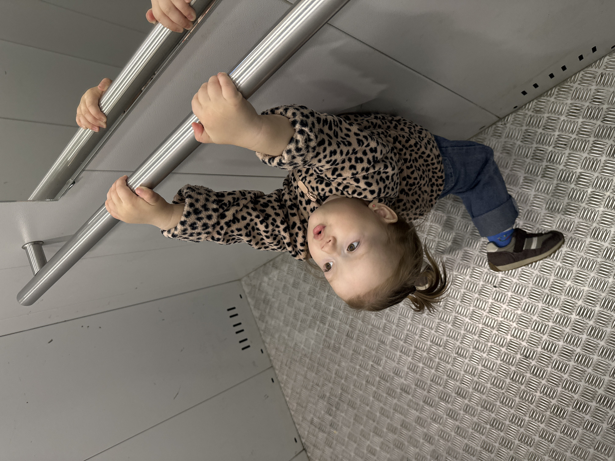 The image captures a young child inside what appears to be a public transport vehicle, such as a subway car or tram, in Yekaterinburg, Russia. The child, a toddler with light brown hair styled in a ponytail, is wearing a tan fleece jacket with a black leopard print, cuffed blue jeans, and dark shoes with a white stripe. A small pink stud earring is visible in the child's left ear. The child is holding onto a horizontal silver metal handrail with both hands, with their body leaning against it, and gazing upwards and to the right with a slightly open mouth.
The interior environment features light grey, possibly metallic, wall panels with visible seams. On the upper left wall, there are dark rectangular perforations arranged in groups, suggestive of vents or fasteners. More small black square perforations are visible along the base of the wall on the lower right. The floor is constructed from a silver-colored textured metal plate, exhibiting a diamond or checker plate pattern. A mirror on the right wall reflects the handrail and the child's hands, creating a duplicated visual effect.