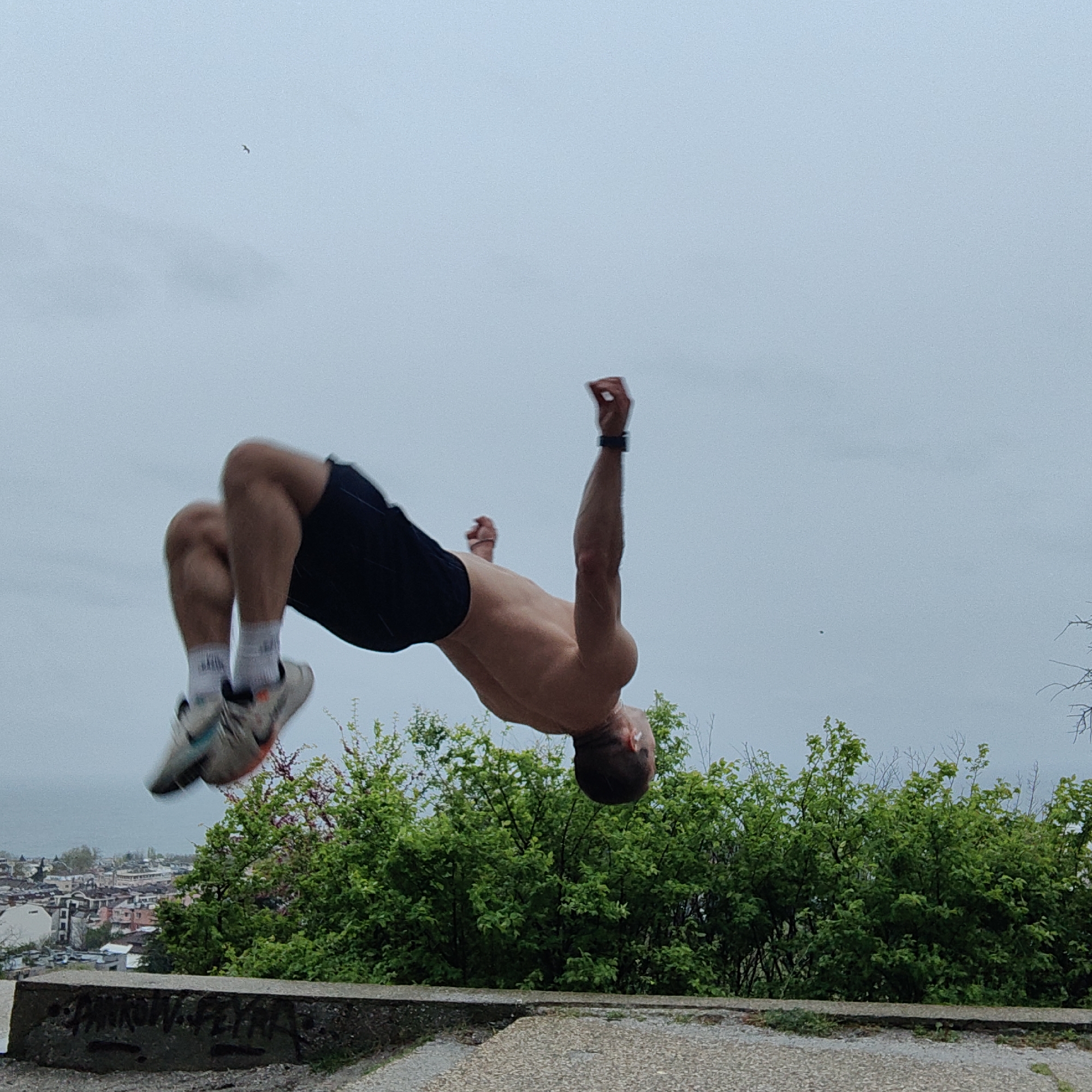 The image depicts a male individual performing an athletic backflip outdoors against an overcast sky. He is shirtless, displaying an athletic build, and wears dark shorts, white socks, and light-colored athletic shoes with visible blue and orange accents. A dark watch or wristband is visible on his left wrist. He is captured mid-air, with his body in a tucked position as he rotates.
The setting is an elevated outdoor location, likely a viewpoint. In the foreground, a rough concrete ledge is visible, covered with some dark, illegible graffiti that appears to say "AMRON_REYA" or similar. Behind this ledge, lush green bushes with dense foliage extend across the mid-ground. Beyond the greenery, a city extends into the distance, featuring numerous buildings under the cloudy sky, consistent with the location context of Varna, Bulgaria. A faint body of water, possibly the Black Sea, can be seen on the distant left horizon. Two small birds are visible flying high in the pale, overcast sky. The overall scene suggests a cloudy daytime, emphasizing the dynamic action against a muted urban and natural backdrop.