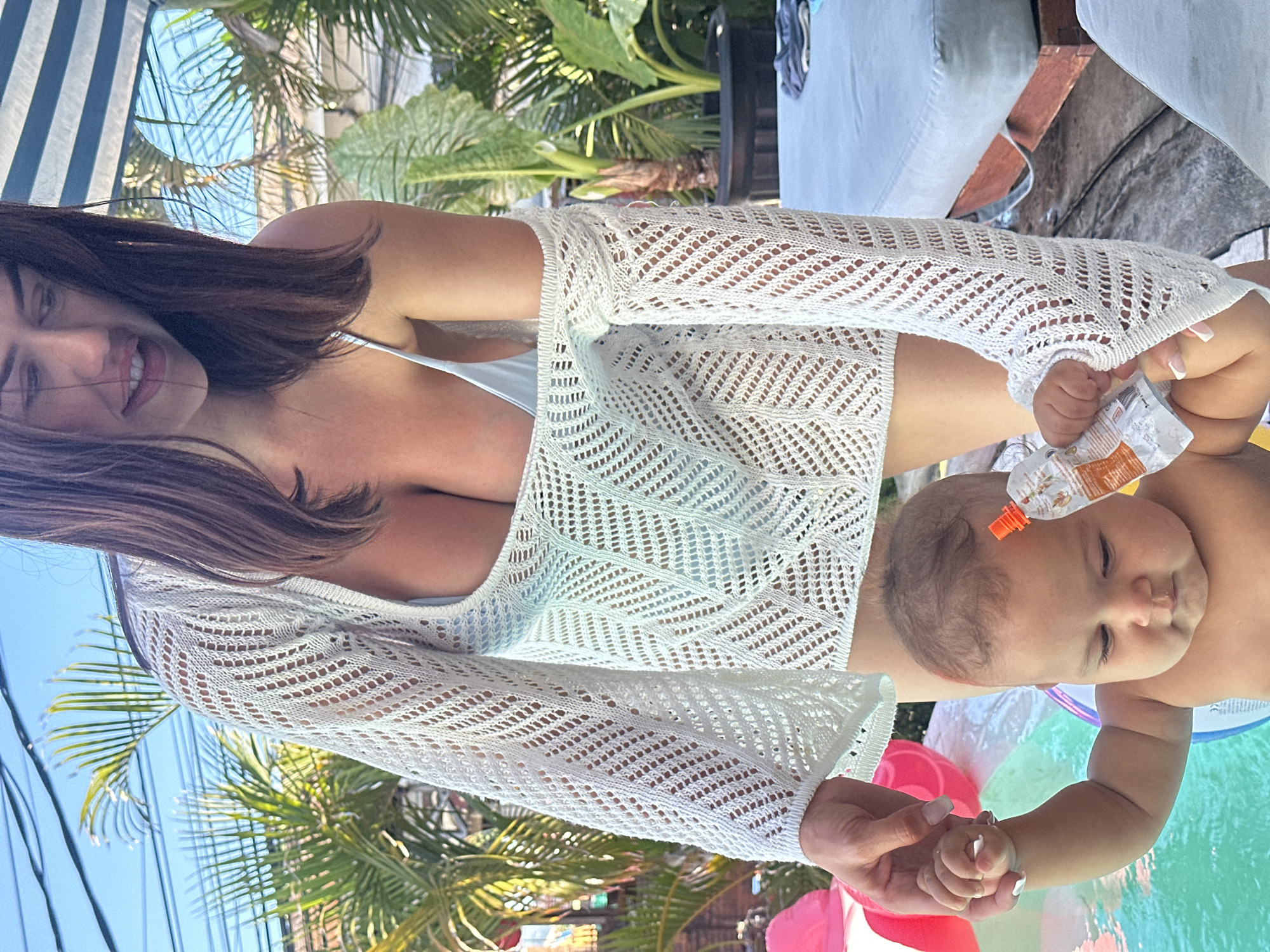 A woman is holding a baby, with an open pouch of food on the baby’s forehead, outdoors on a sunny day. The woman is fair-skinned with long brown hair and is wearing a white crocheted top, with a white bathing suit visible underneath. The baby is also fair-skinned and has some dark hair. The baby's hand is being held by the woman. They are in a shallow inflatable pool with colored inflatable beach balls.
In the background, there are palm trees and other tropical plants, a blue and white striped umbrella, and a table with a light-colored tablecloth. Given the tropical plants, sunny weather, and location of Kuta, Indonesia, it's likely the photo was taken in a backyard or patio setting.