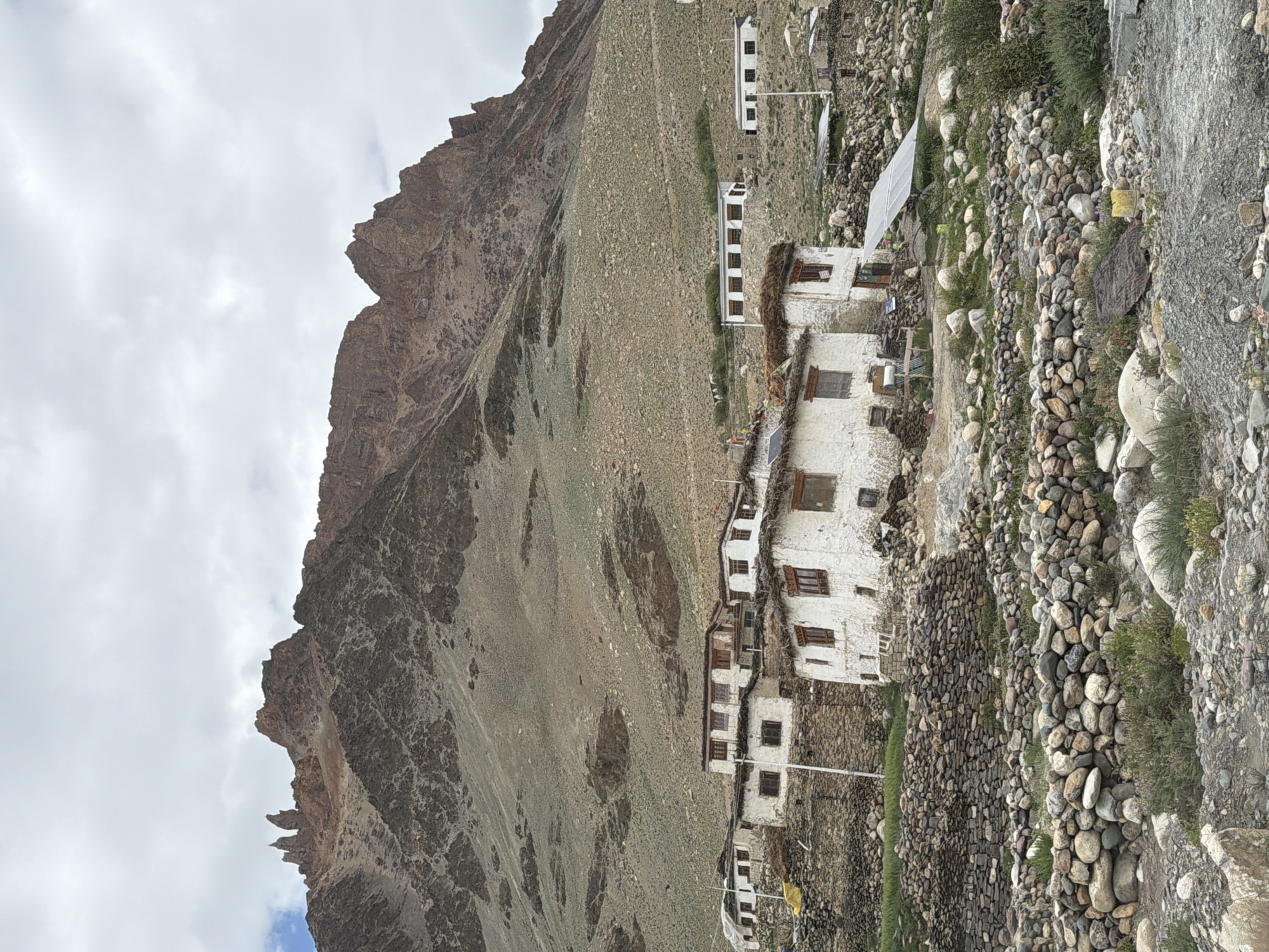 This is a vertical, eye-level, full shot of a remote village nestled at the base of a towering mountain range in Kurgiakh, India. The sky is overcast with heavy, gray clouds. The mountain is a dominant feature, with jagged, rocky peaks in shades of brown and gray, its slopes covered in sparse, dry vegetation and scree.
In the midground, a cluster of traditional white-washed houses with thatched roofs and wooden window frames is situated on a rocky slope. The houses are built with stone walls and plaster. Some of them appear to have solar panels installed. The foreground is a rugged terrain of loose rocks and pebbles, interspersed with dry grasses and small shrubs. There are no people or animals visible in the scene. The overall impression is one of a harsh, remote, and sparsely populated environment.