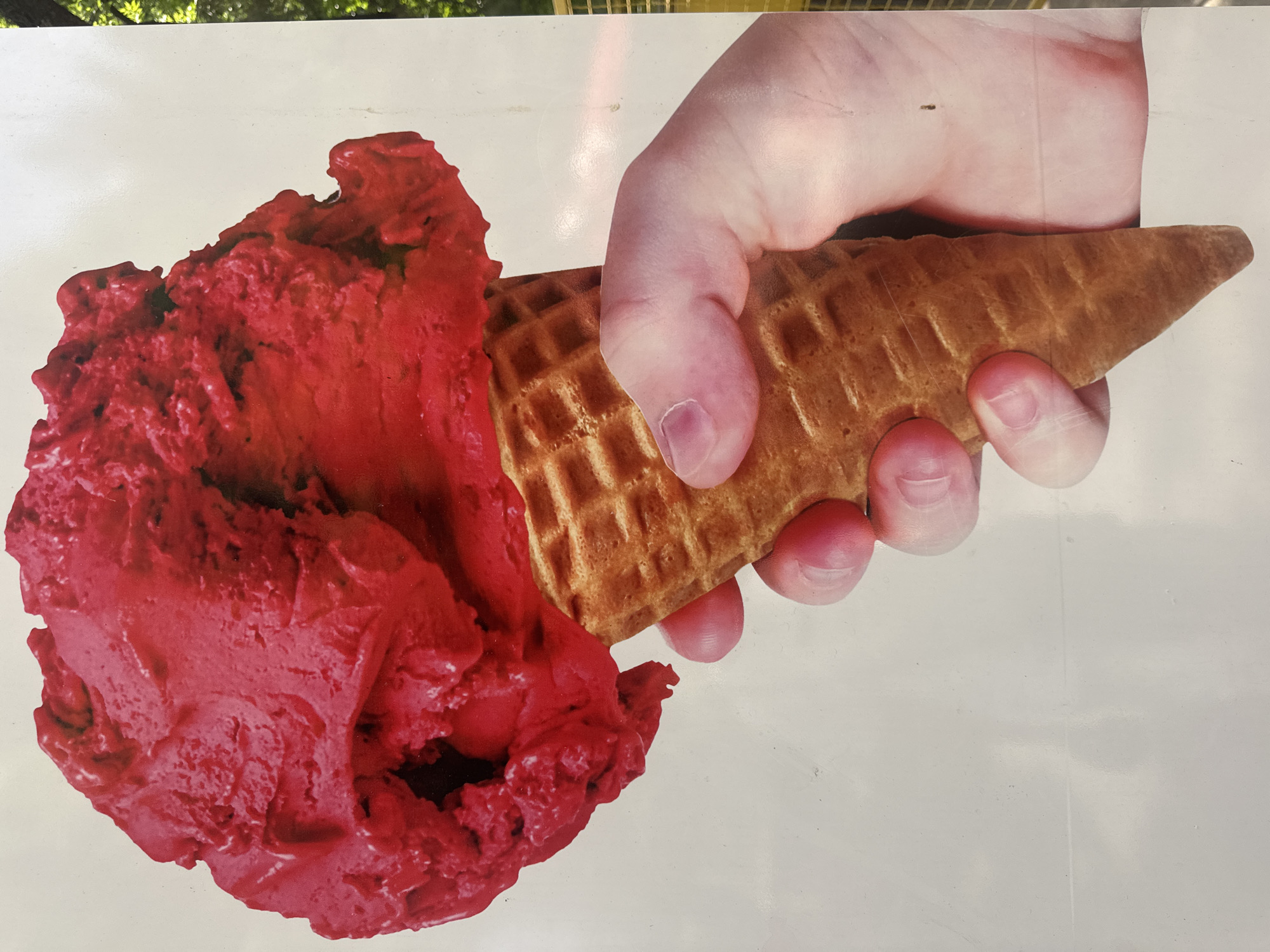 A close-up shot of a hand holding a red ice cream cone against a white background. The ice cream appears to be a vibrant red, possibly strawberry or raspberry flavored, with visible scoops and swirls. The cone is a golden-brown waffle cone, with its characteristic grid pattern. The hand is light-skinned, with pinkish fingernails. The background is predominantly white, with some subtle shadows and light reflections. In the upper right corner, a blurred glimpse of green foliage suggests an outdoor setting, possibly trees or bushes. The overall impression is of a refreshing treat on a bright day. This image has no discernible location in Tashkent, Uzbekistan.