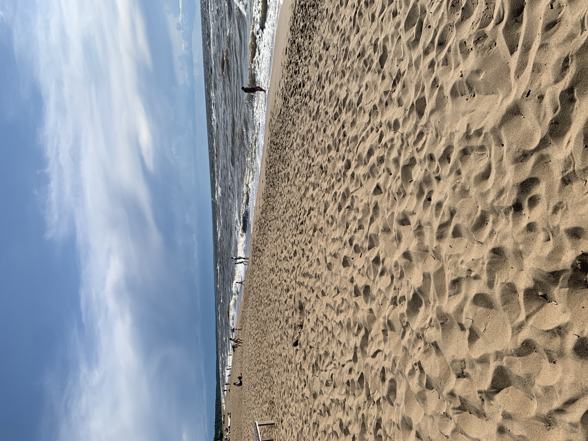 This is a medium-angle shot of a sandy beach in Saulkrasti, Latvia. The foreground is dominated by textured sand with footprints and small debris. In the middle ground, a few people are scattered along the shoreline. Some are walking, others are sitting or playing near the water. The sea is choppy with moderate waves breaking on the shore. The background shows a tree line, indicating the edge of the land. Above, a partly cloudy sky with dramatic, wispy clouds fills the upper portion of the image. The lighting suggests it is daytime, possibly late afternoon due to the soft quality of light.